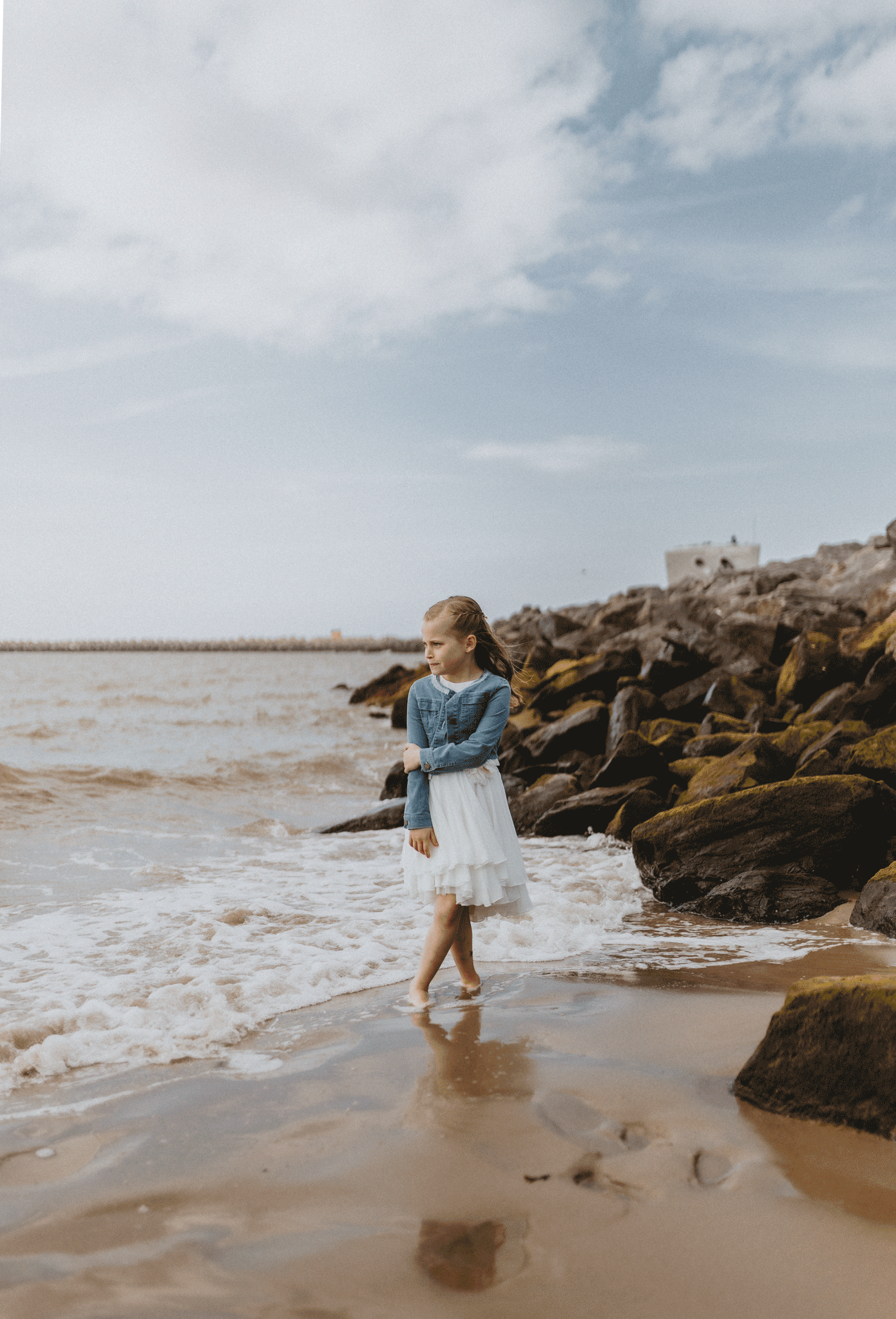 Meisje in een witte jurk staat op rotsen bij het strand, met de golven die zachtjes tegen de stenen slaan op het strand van Oostende