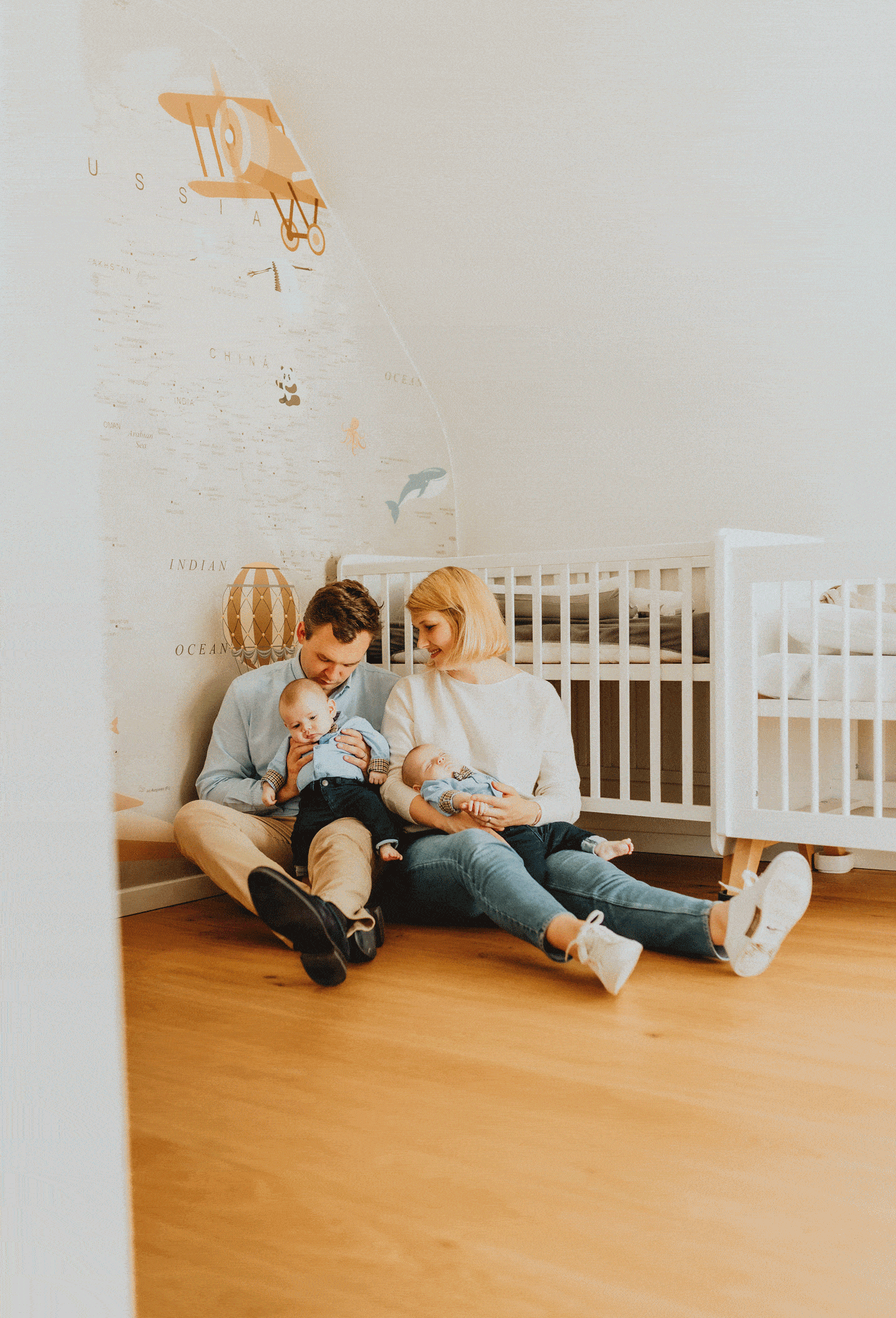 Een koppel en hun tweeling baby's zitten op de grond in een kamer met een muurschildering van een wereldkaart
