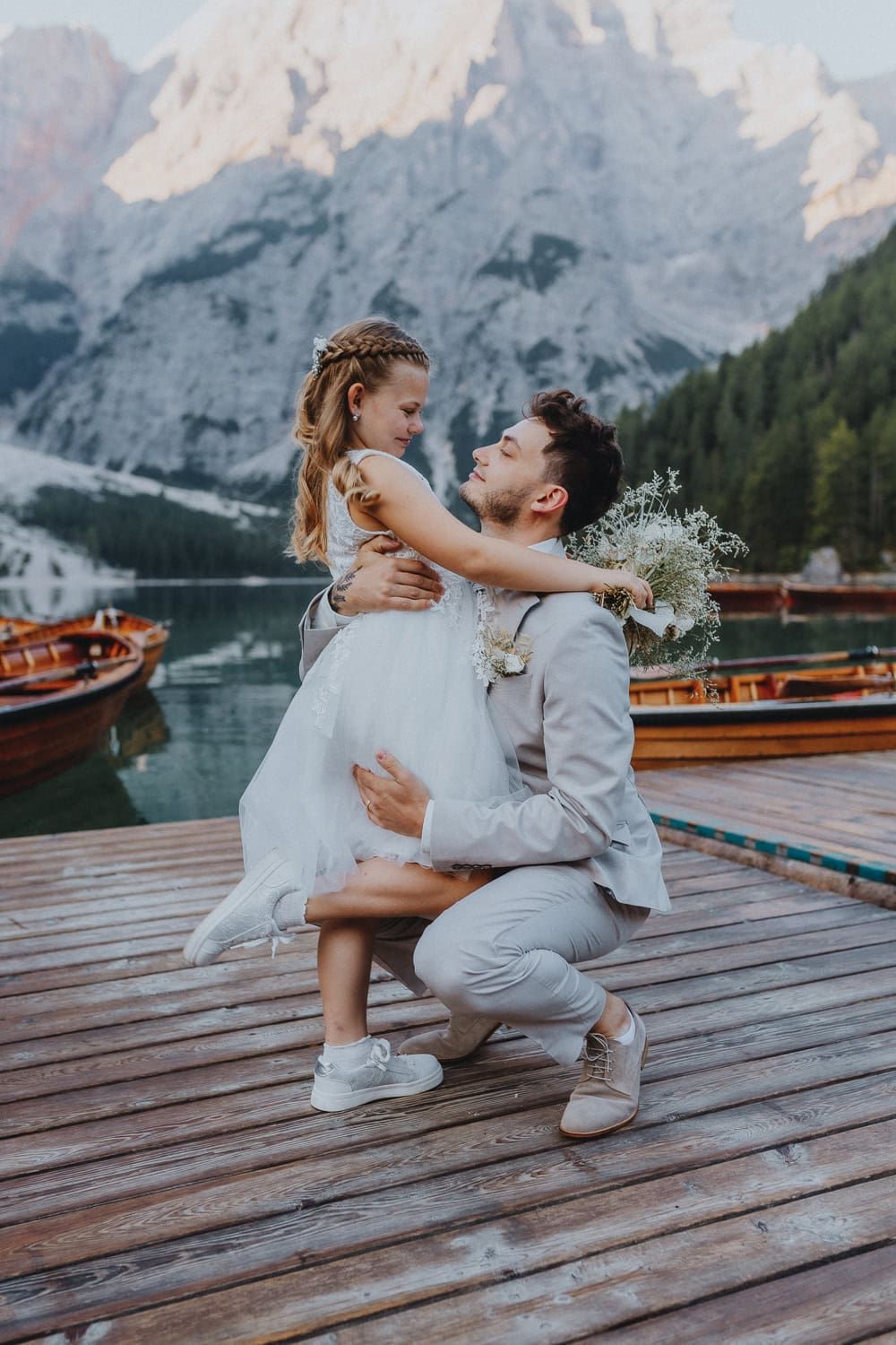 Groom kneels on a wooden dock embracing bride with mountain lake and wooden boats behind them.