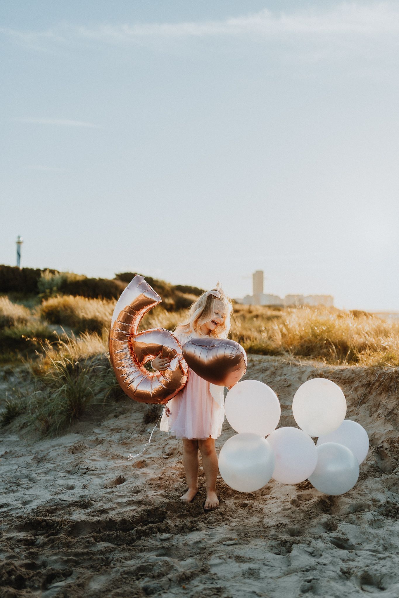 Een meisje in een roze jurk houdt blij een groot cijfer 6-ballon vast op een zonnig strand in West-Vlaanderen. Hartballon erbij.