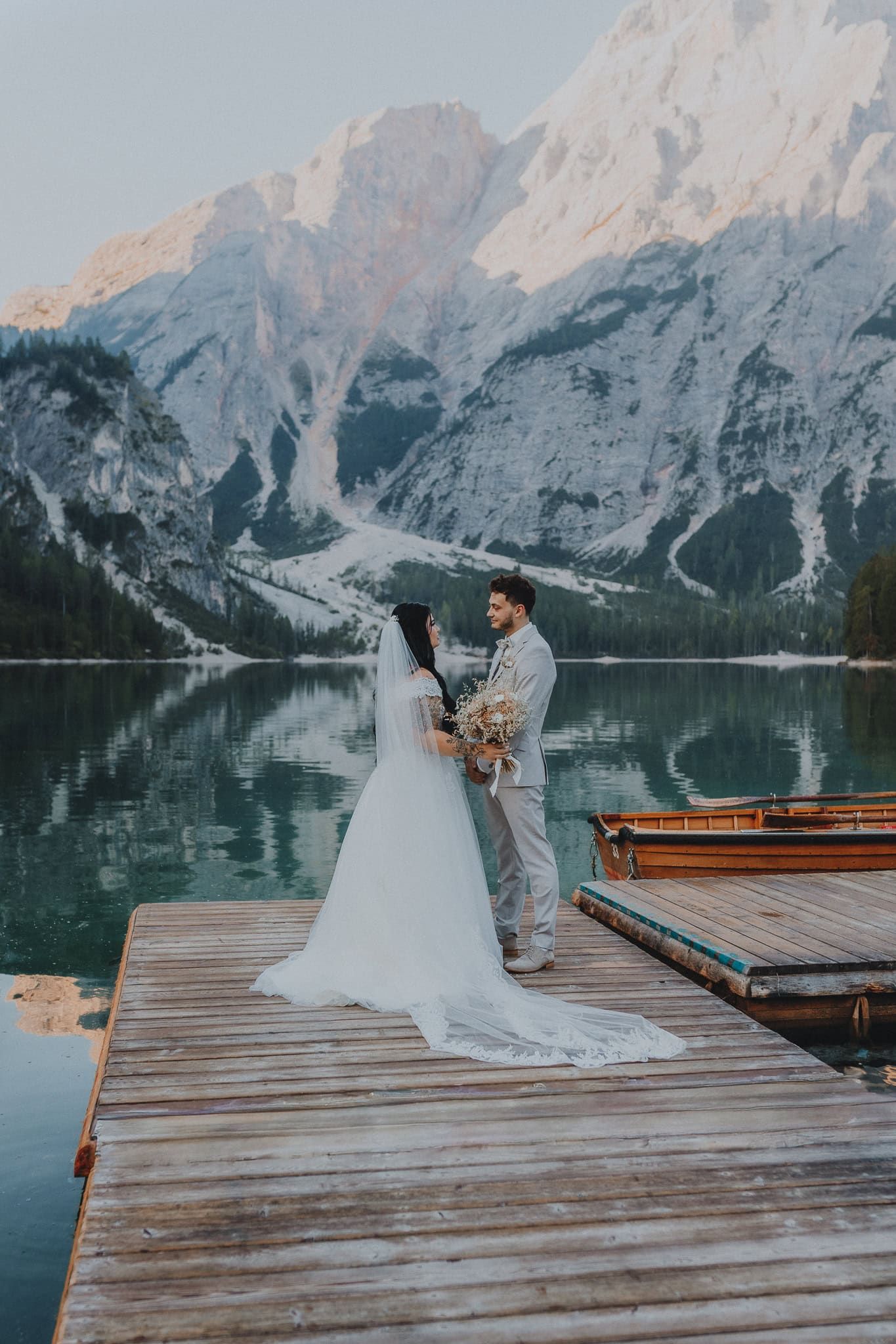 Bride and groom standing on a wooden dock with stunning mountain lake reflection at sunset.