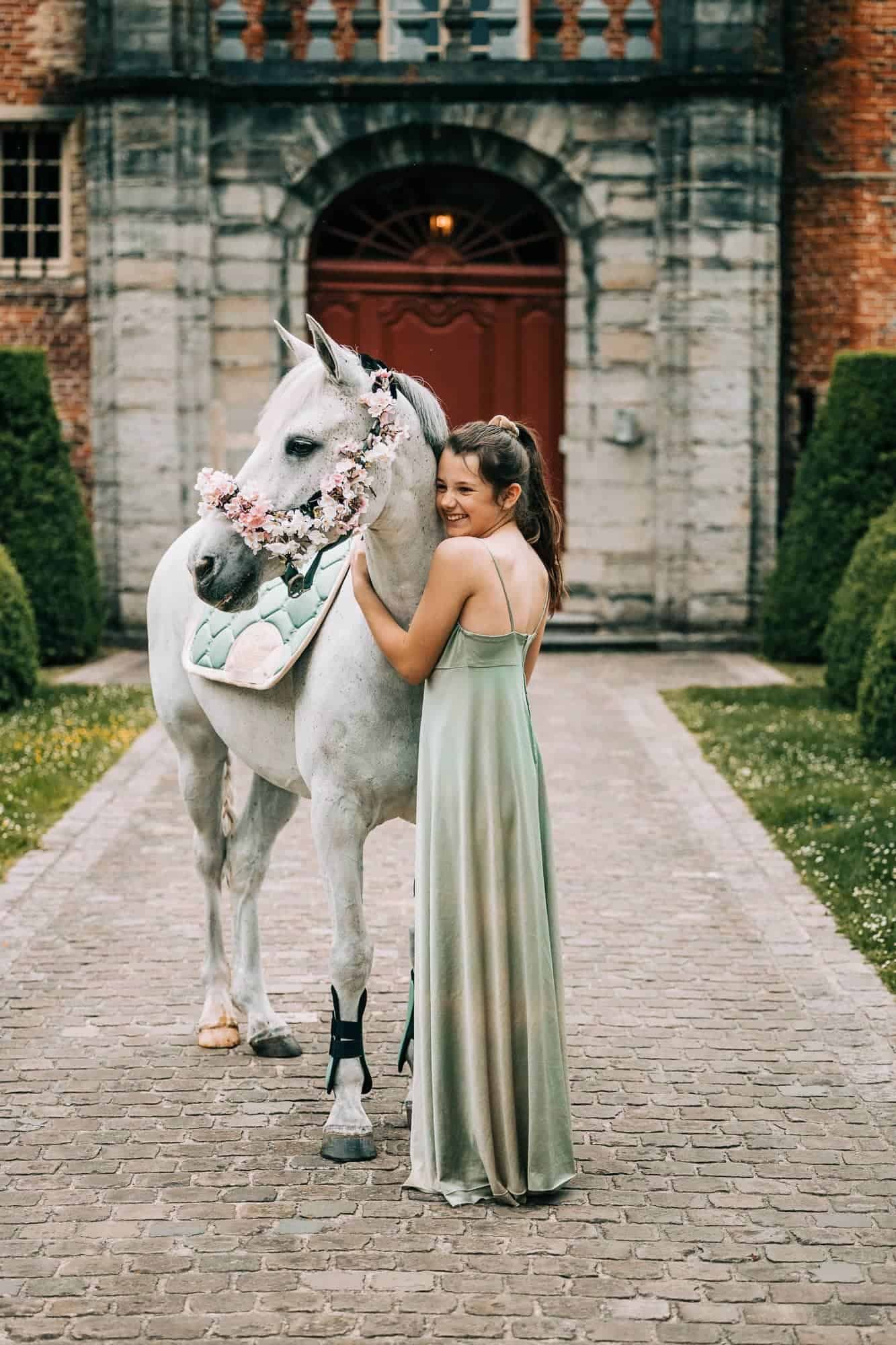 Meisje in groene jurk omhelst wit paard met bloemenkrans op stenen pad in Roeselare. Elegant en vrolijk moment tijdens communie shoot.
