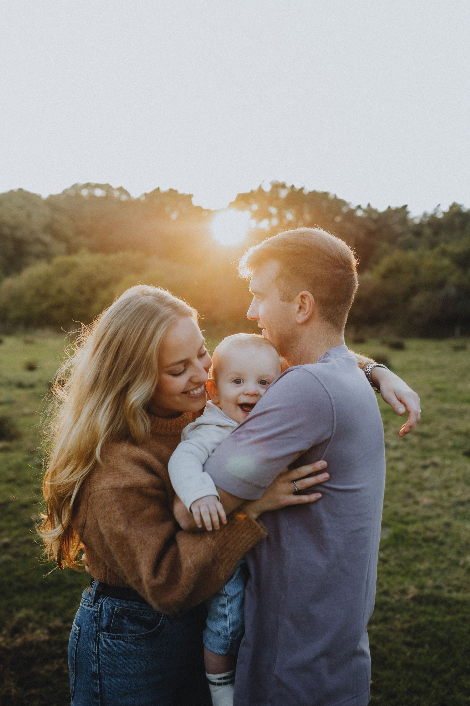Young parents holding a laughing baby in a sunlit green field at golden hour.