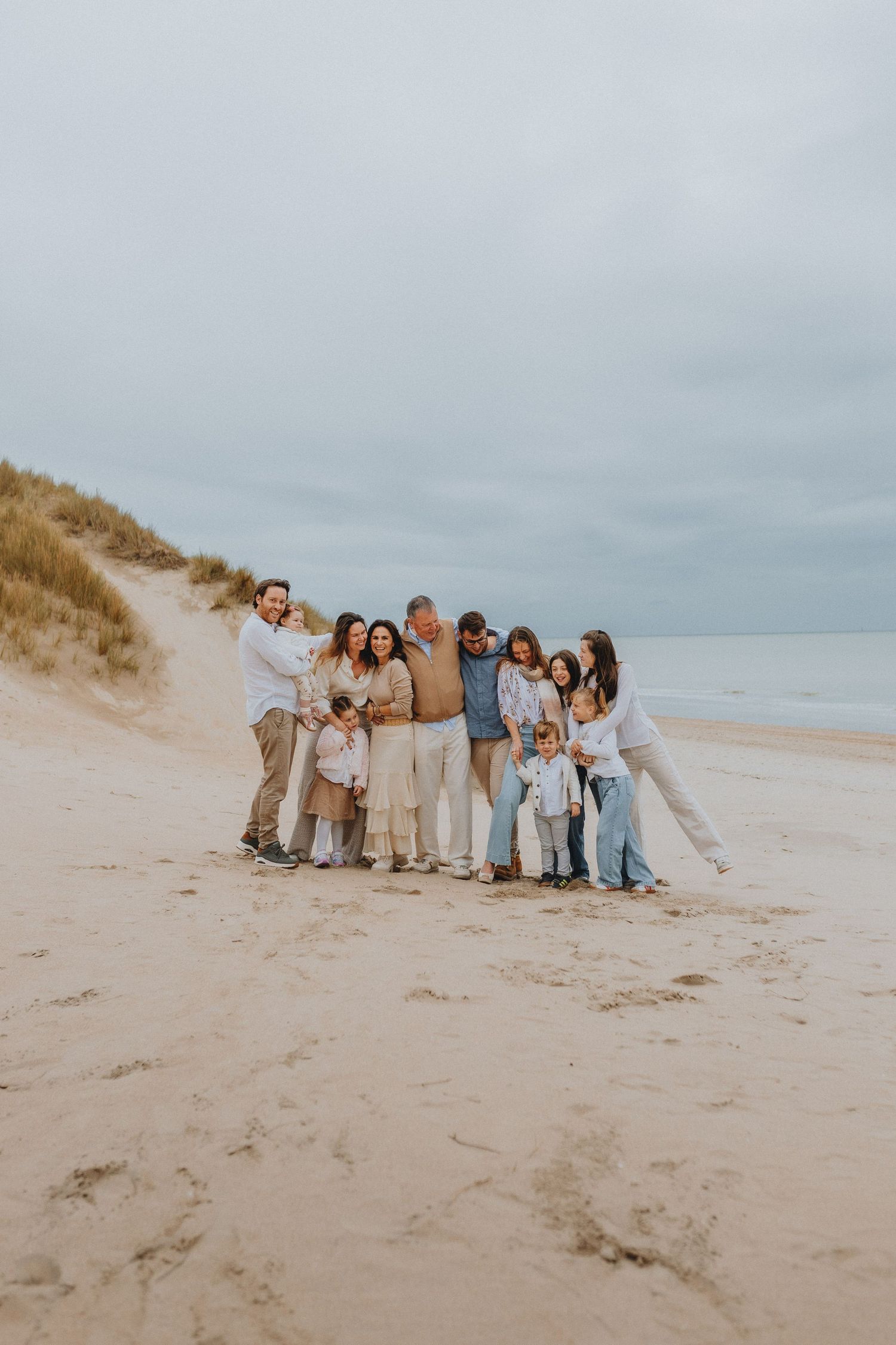 Large extended family group of eleven posing together on a wide sandy beach under cloudy skies.