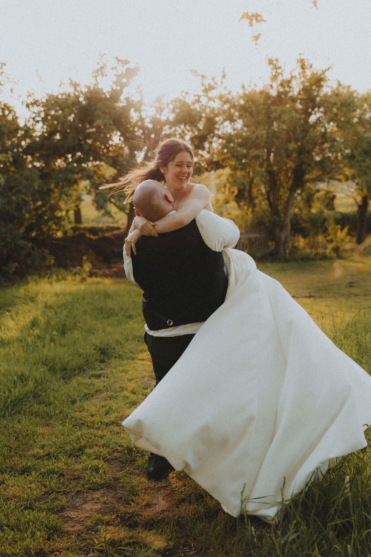 Groom sweeping bride in white gown off her feet outdoors at golden hour in a lush green garden setting.