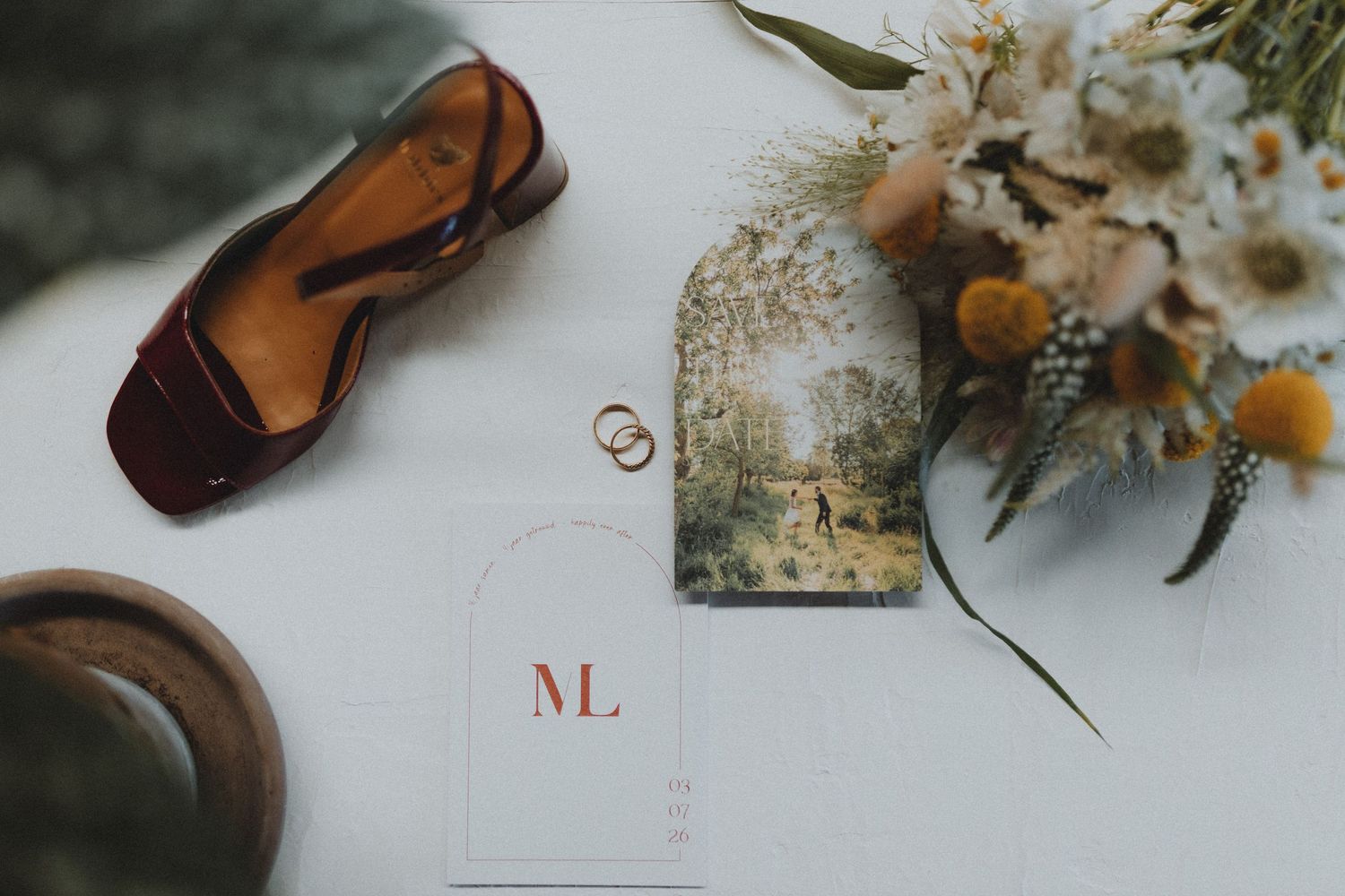 Flat lay of wedding details including rings, floral bouquet, monogram card, sunglasses, and wooden bowl on white surface.