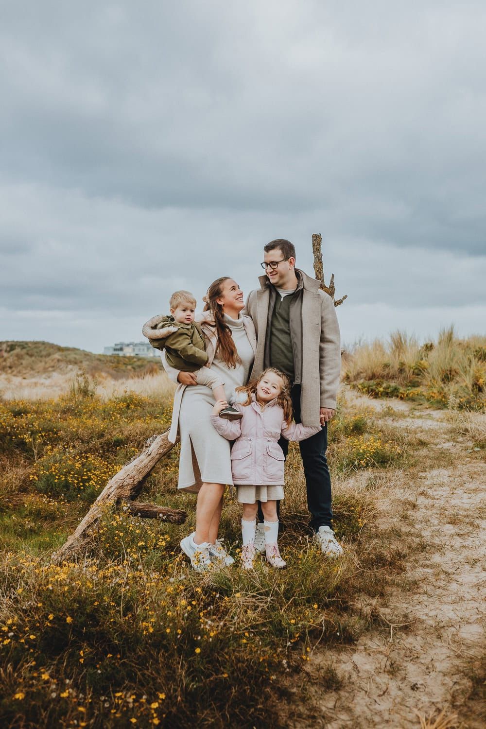 Een vrolijk gezin staat buiten op een bewolkte dag. Ouders houden de handen vast van een jongen en meisje op een pad met gras en wilde bloem