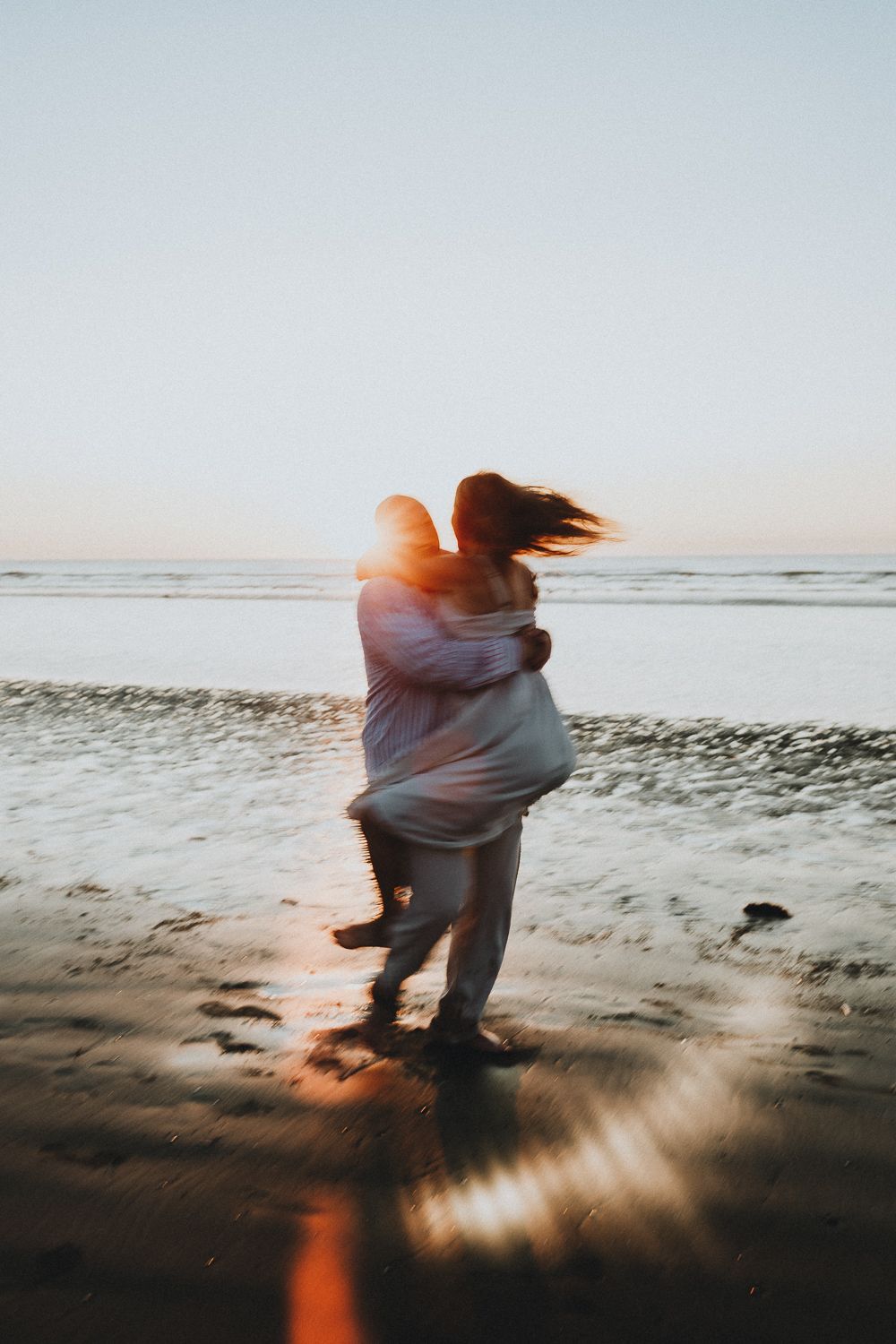 Man lifting woman in his arms on a wet beach at sunset, motion blur capturing joyful movement.