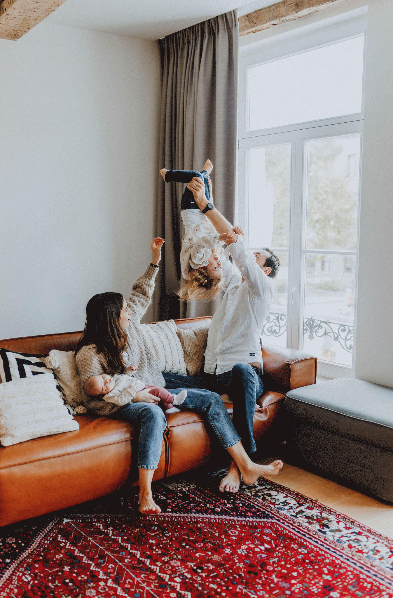 Vrolijk gezin in een gezellige woonkamer. Man tilt kind speels ondersteboven, vrouw glimlacht met baby op leren bank.