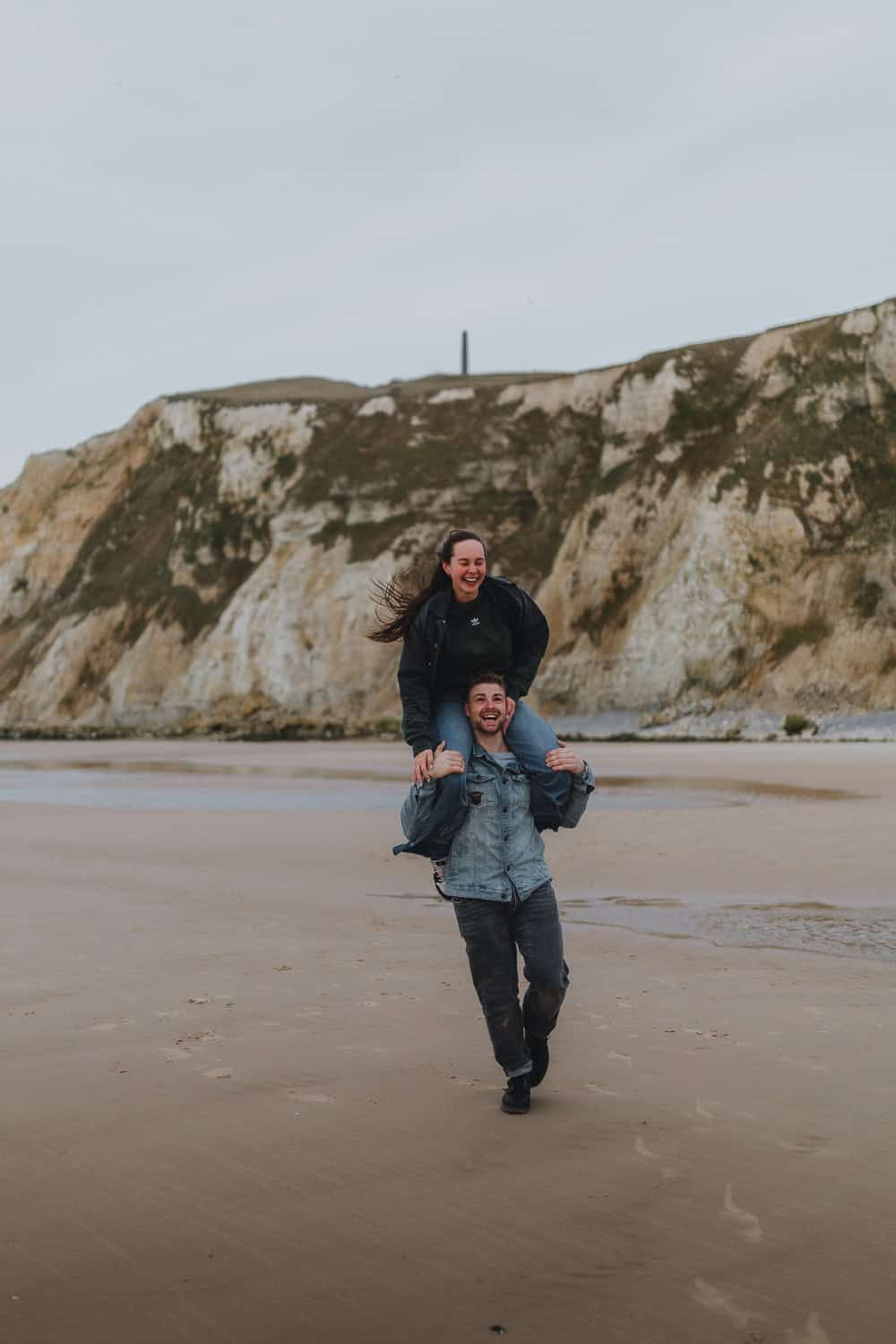 Couple laughing on a sandy beach, man carrying woman on shoulders with white cliffs in the background.