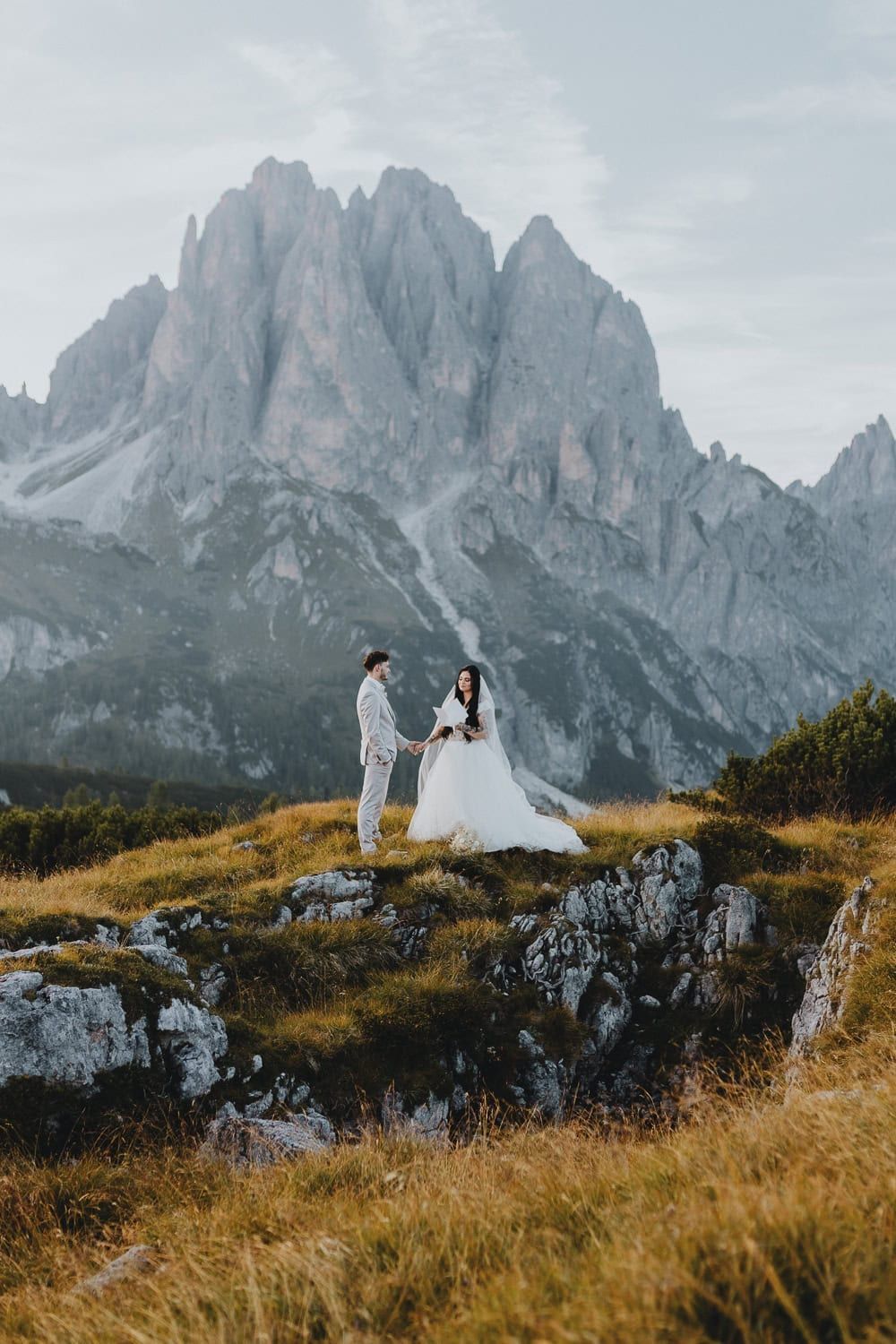 Elegant bride and groom standing on rocky alpine terrain with towering Dolomites mountain peaks behind them.