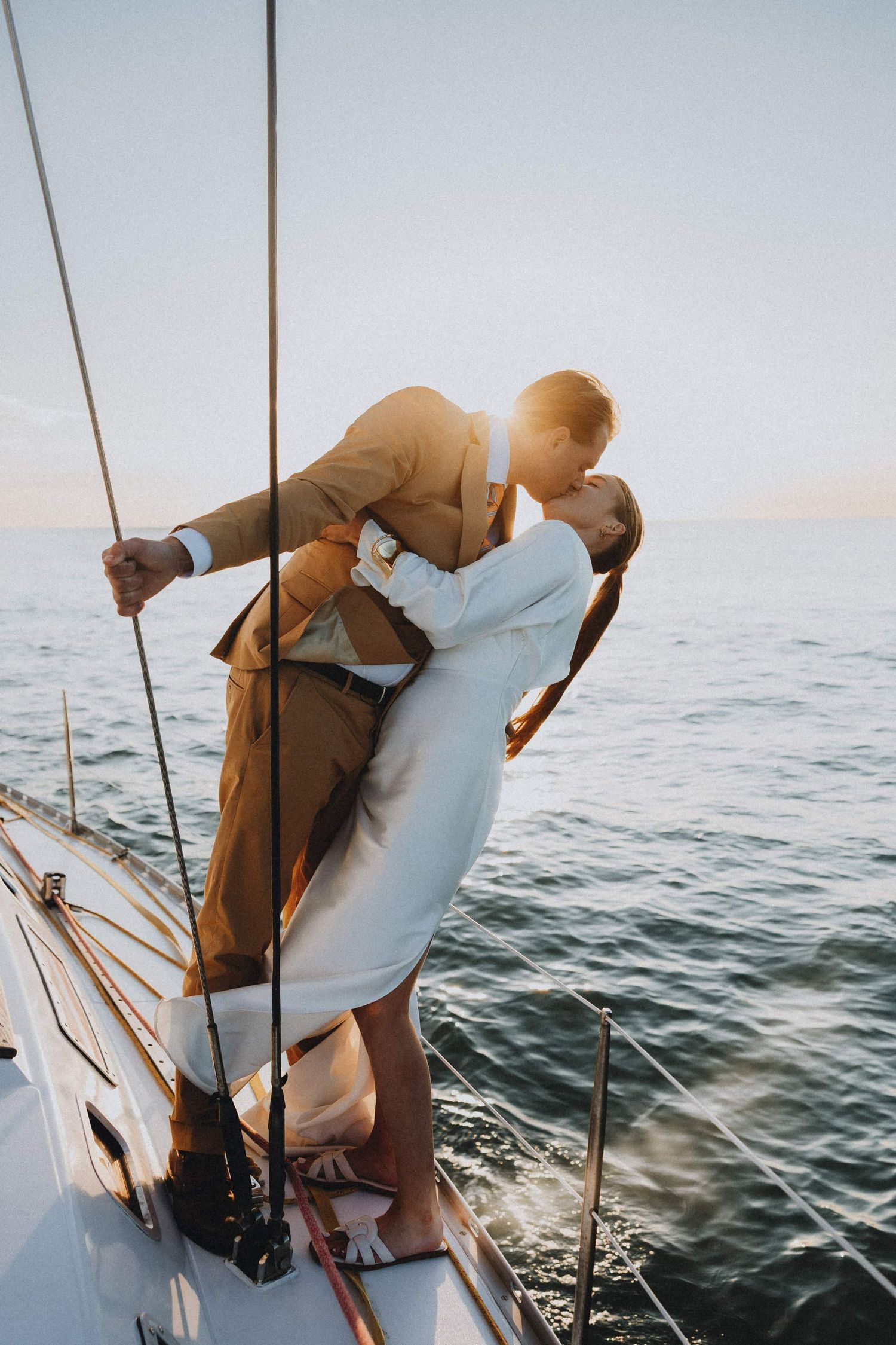 A couple shares a romantic kiss on a sailboat at sunset, with the calm ocean stretching behind them.