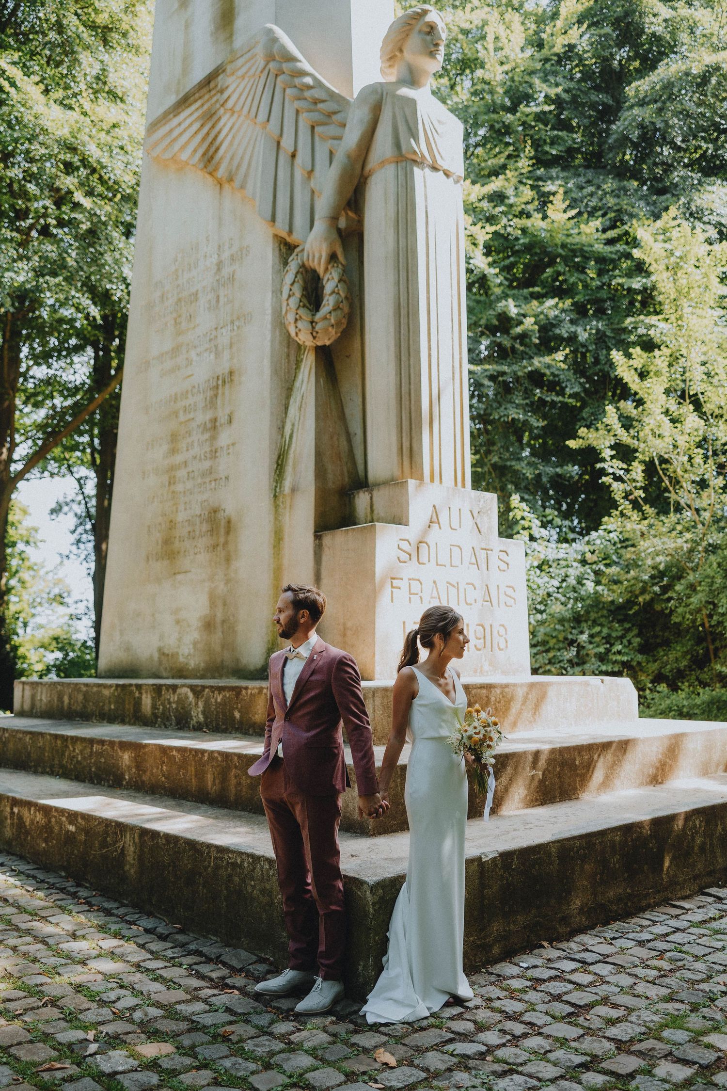Bride and groom holding hands before a French war memorial monument with angel sculpture outdoors.