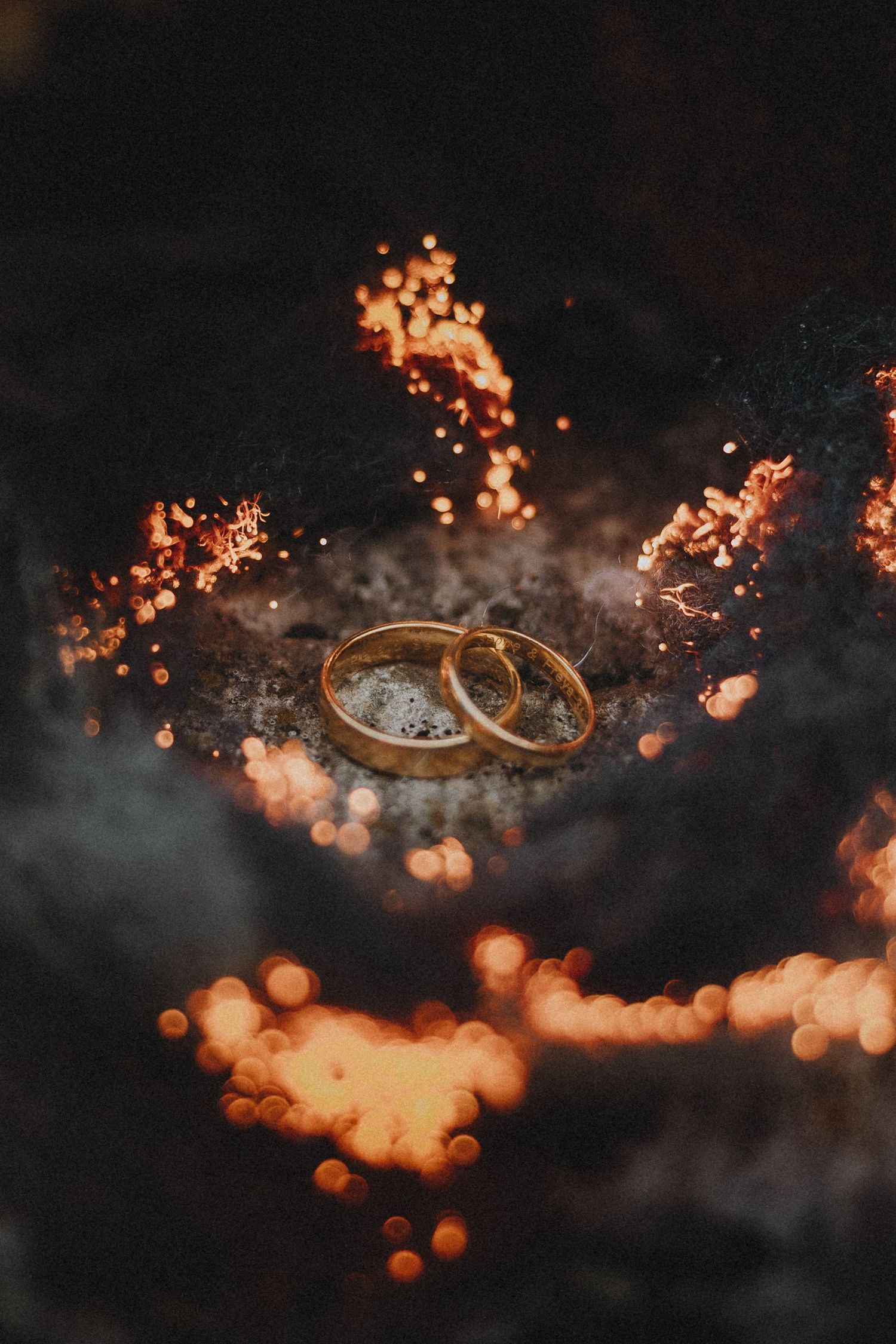 Two gold wedding rings resting on dark embers surrounded by glowing bokeh lights and sparks.