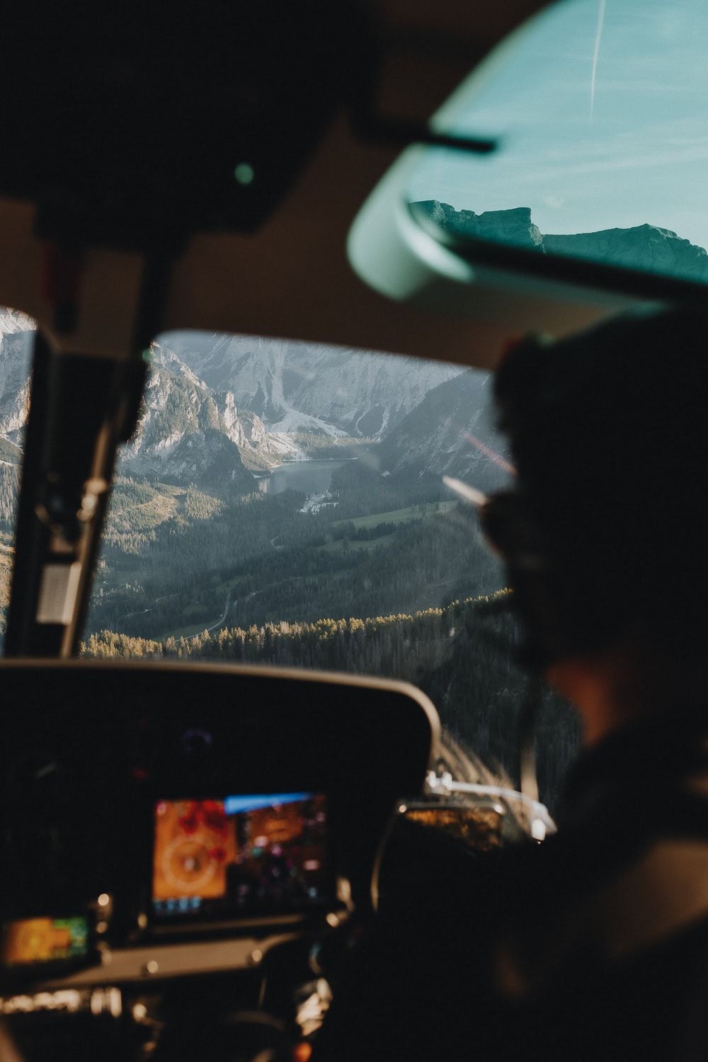 Pilot flying helicopter over stunning mountain landscape with rocky peaks and forest valley visible through cockpit window.