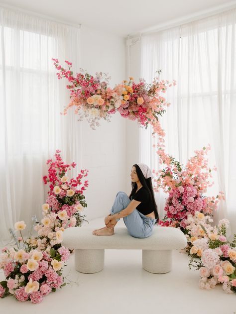 Woman sitting on white bench surrounded by stunning pink and peach floral arrangements in bright studio.