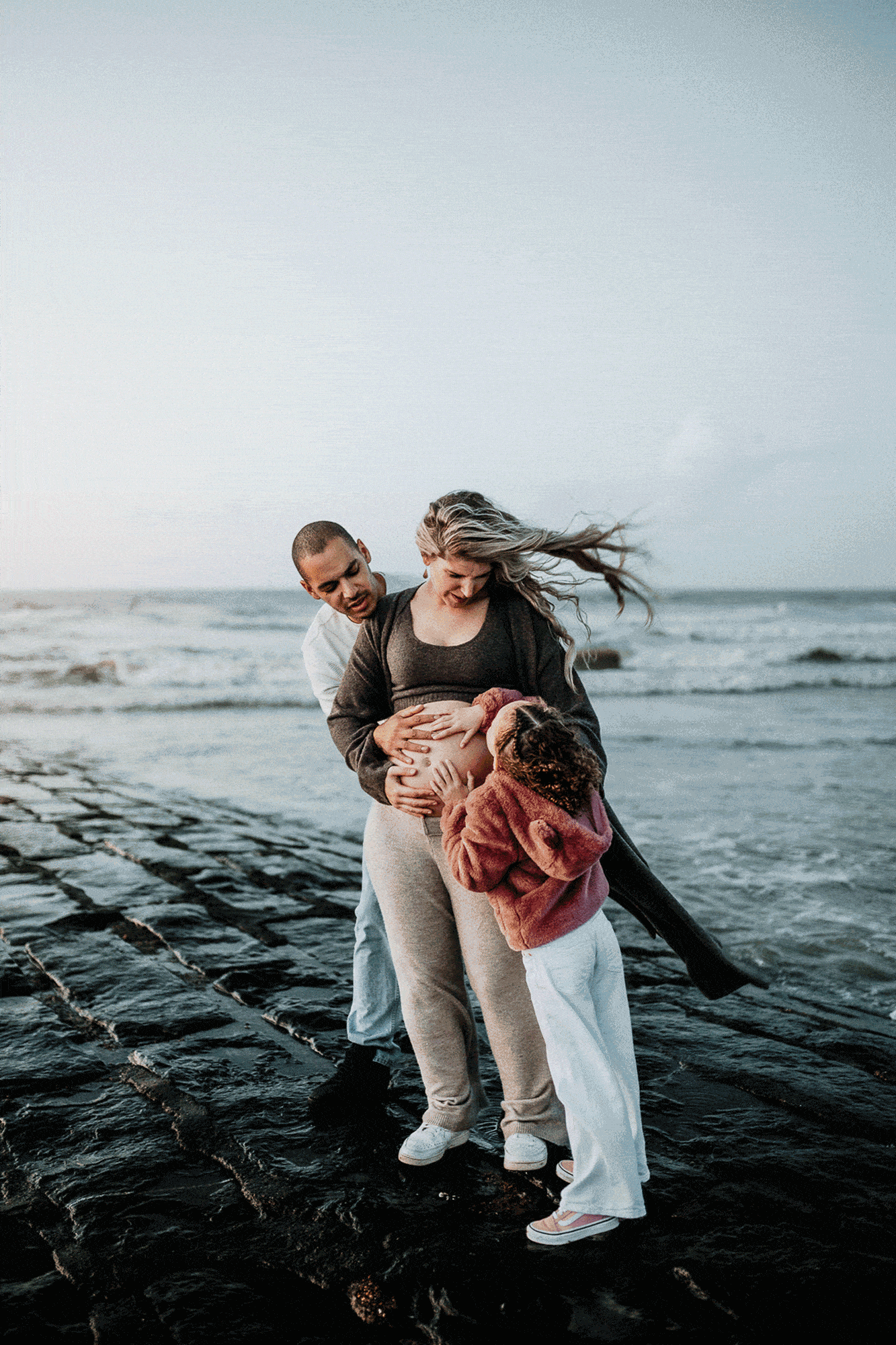 Een zwangere vrouw omringd door haar gezin op het strand, met golven en zand in de achtergrond