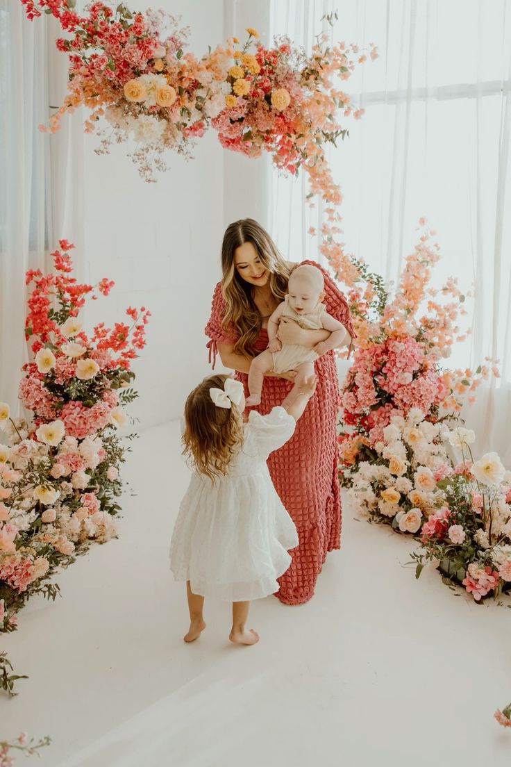Mother in red dress holds baby while young daughter in white dress reaches up, surrounded by pink flowers.