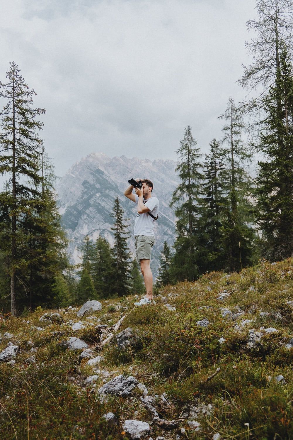 Photographer shooting with camera on rocky hillside surrounded by pine trees and mountain scenery.