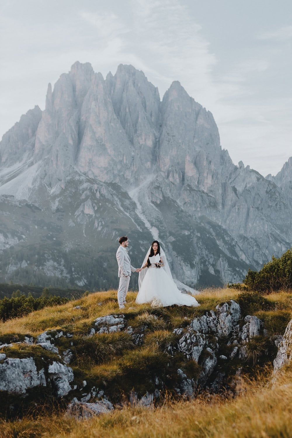 Bride and groom stand holding hands on a grassy hillside with towering Dolomites rocky peaks behind.