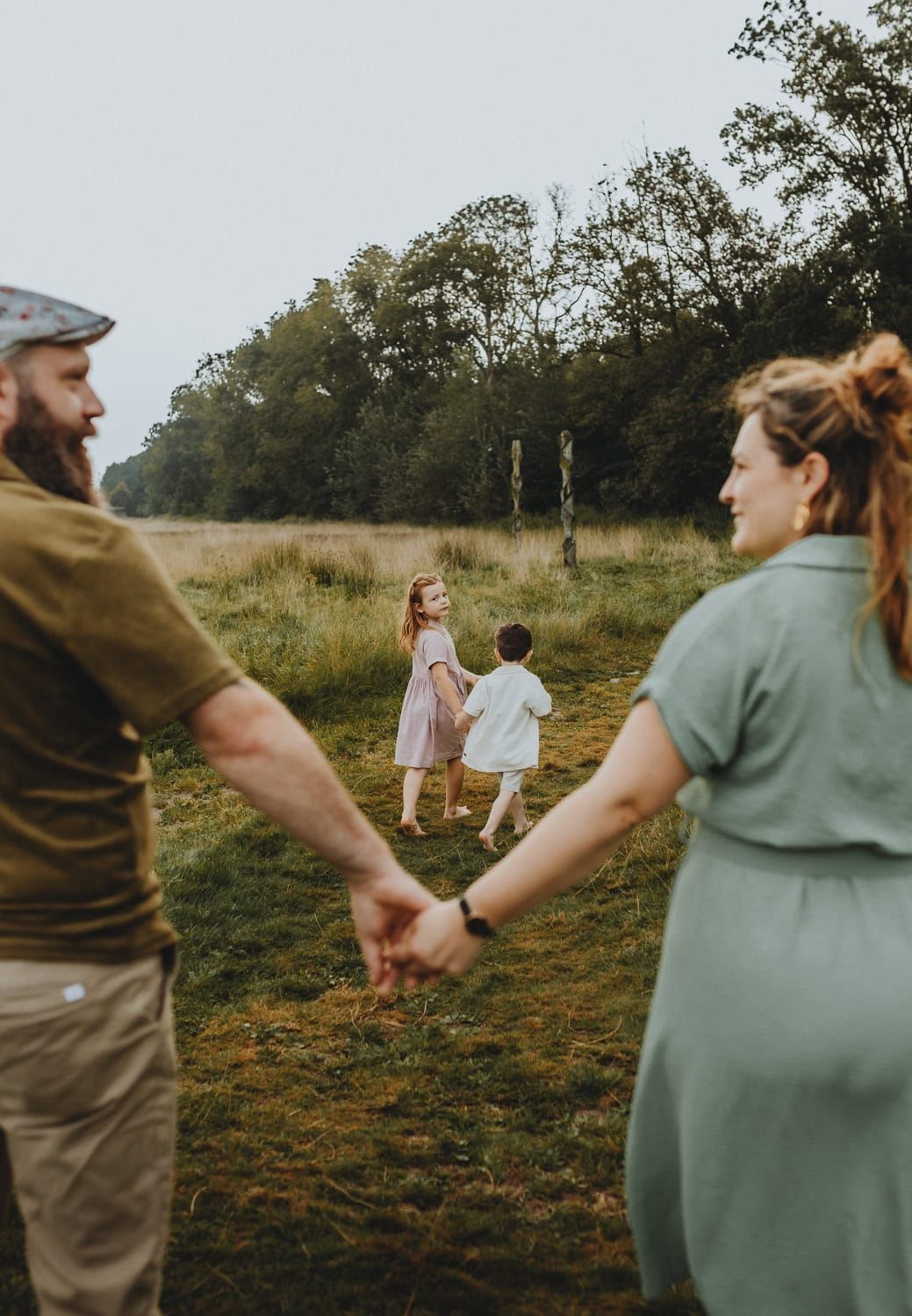 Een gezin houdt elkaars handen vast en wandelt op een grasrijk pad. Ouders lachen, terwijl twee kinderen vooruit lopen en achterom kijken.