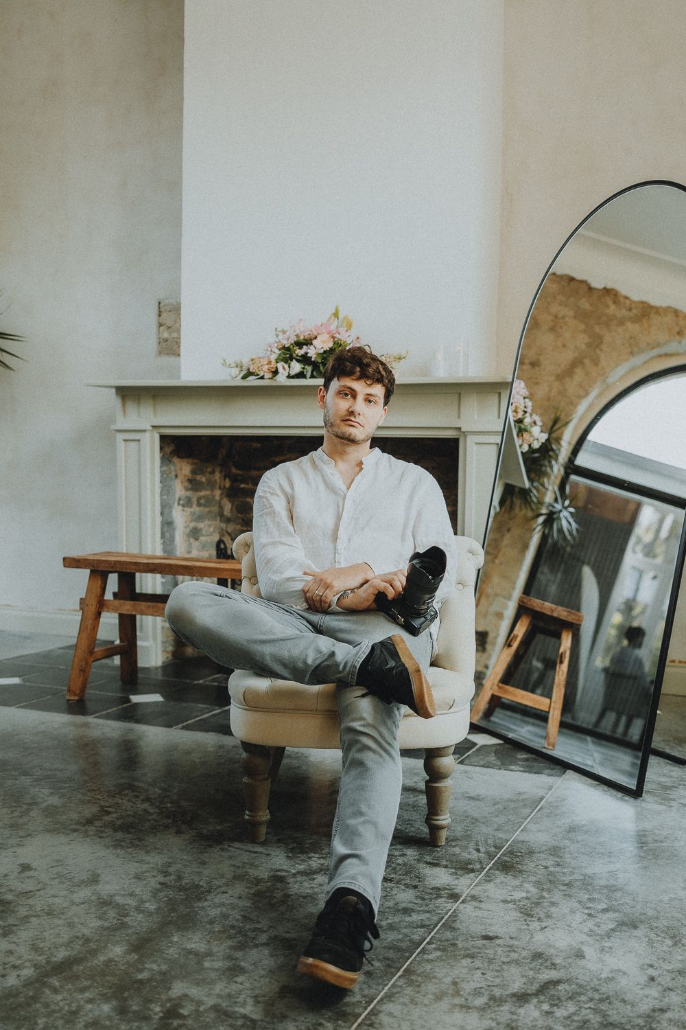 Man in white shirt and gray pants sitting relaxed in armchair near fireplace with floral arrangement and mirror.