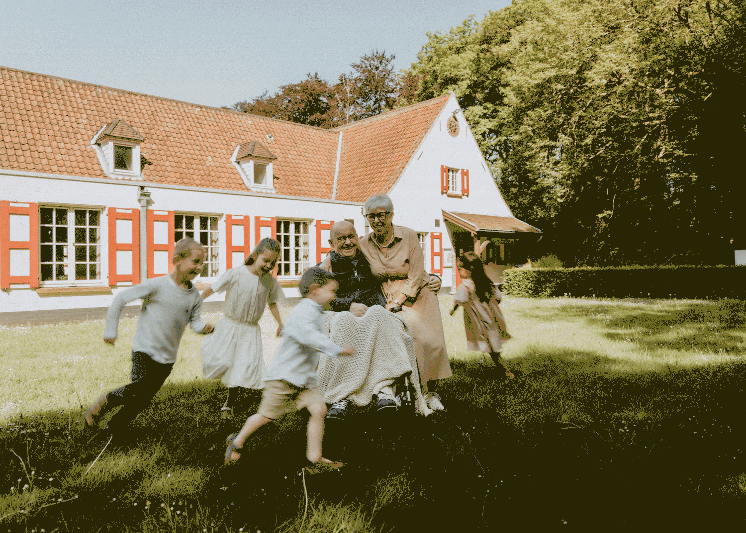 Een vrolijk moment voor een huis met rode luiken; een ouder koppel zit tevreden terwijl kinderen in de zon spelen en rennen.
