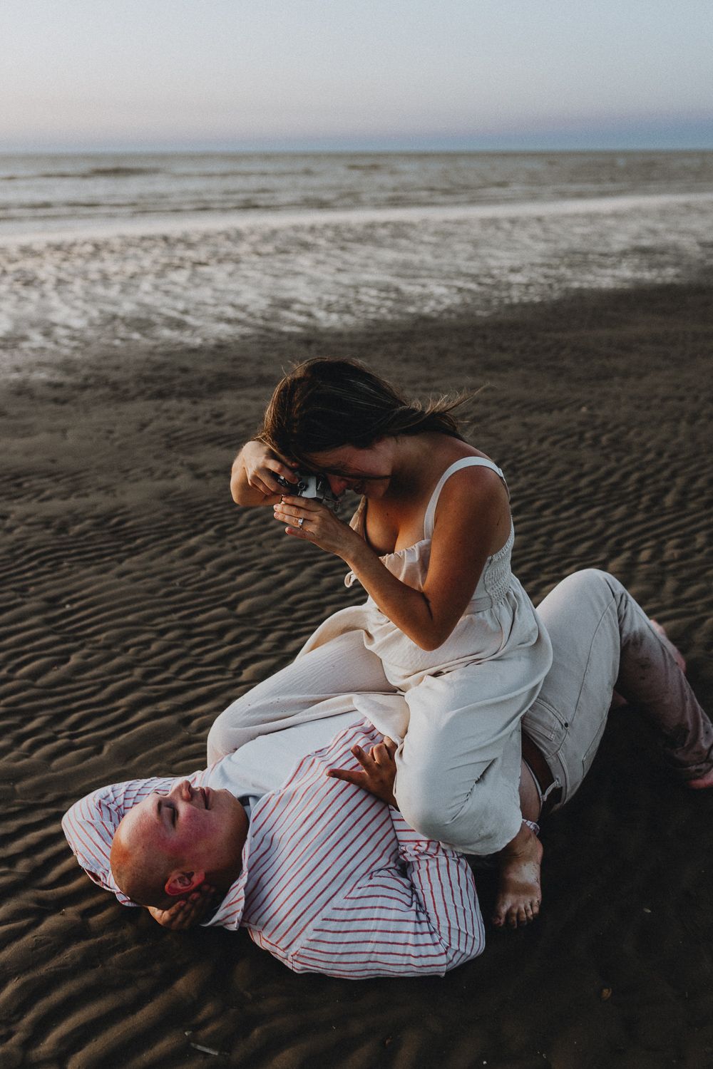 Een vrouw in witte jurk maakt speels foto terwijl ze op de borst van een man zit op het strand. Hij ligt lachend in het zand.