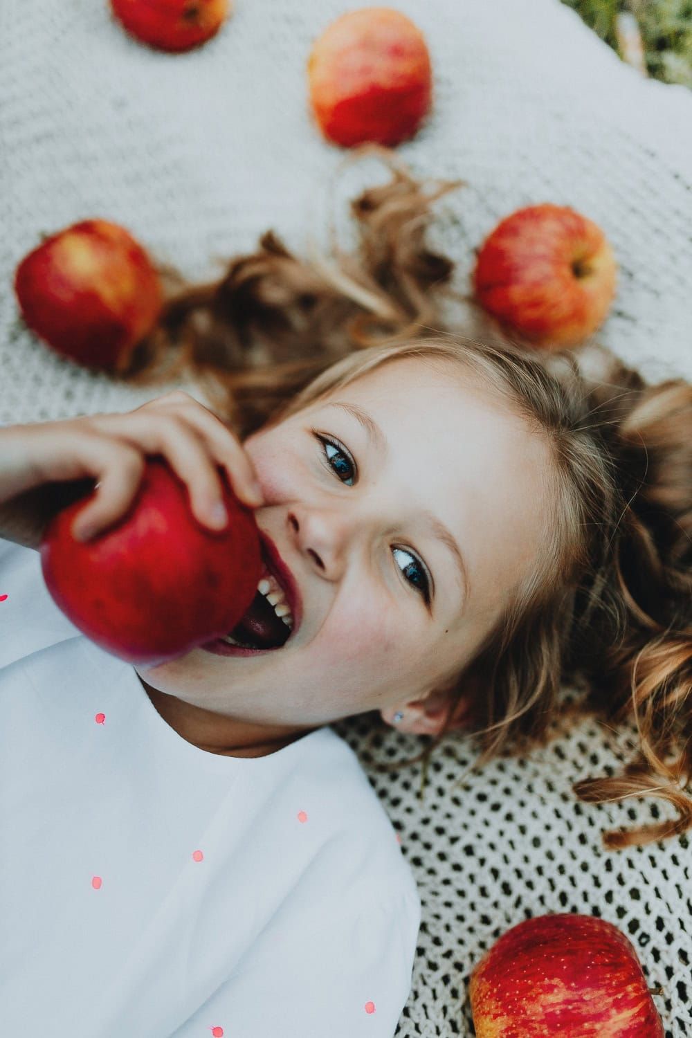 Een lachend kind ligt op een deken met rode appels verspreid. Ze bijt vrolijk in een appel. Speels en blij moment tijdens een communie shoot