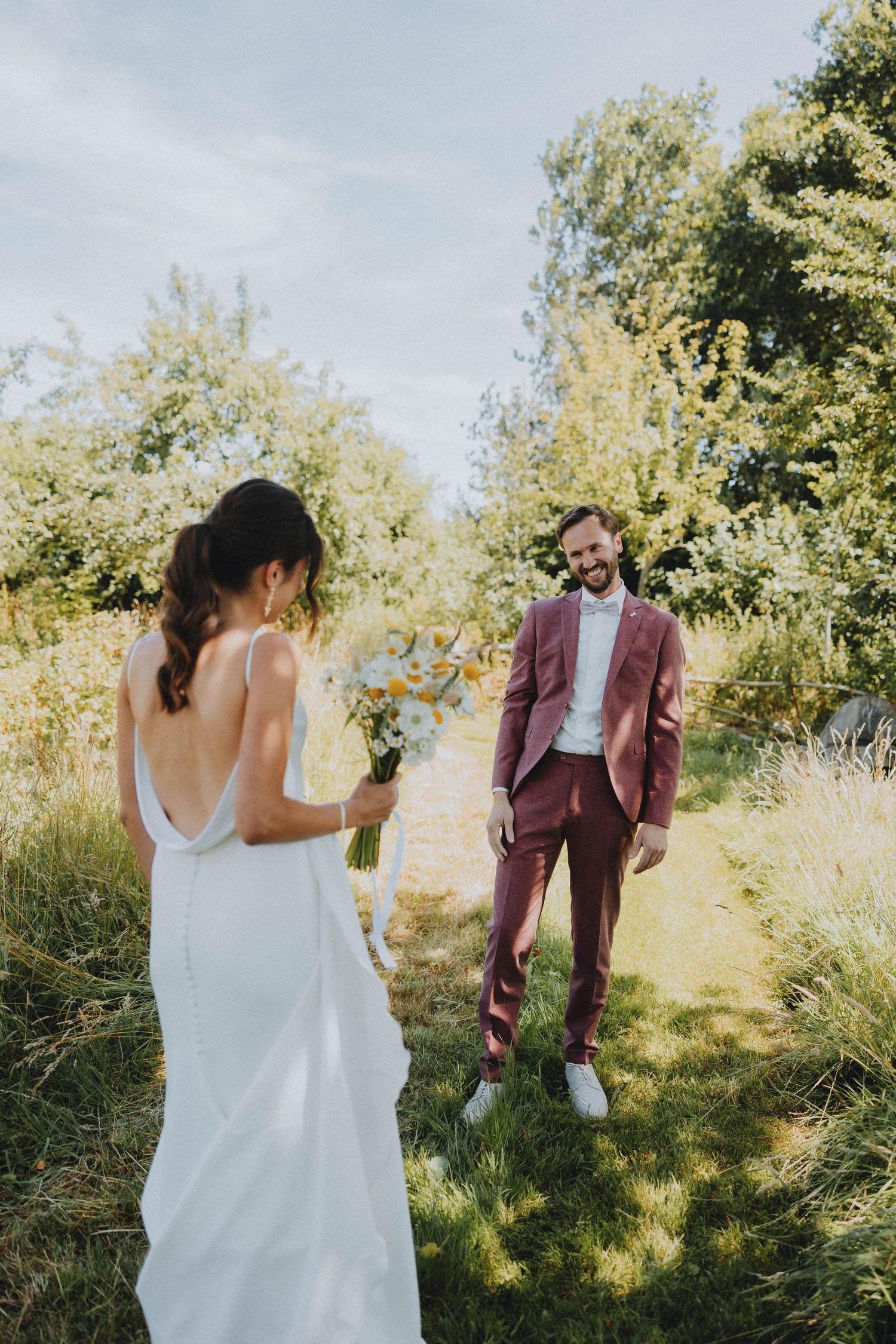Bride in white backless gown holds wildflower bouquet as smiling groom in burgundy suit stands in sunny garden.