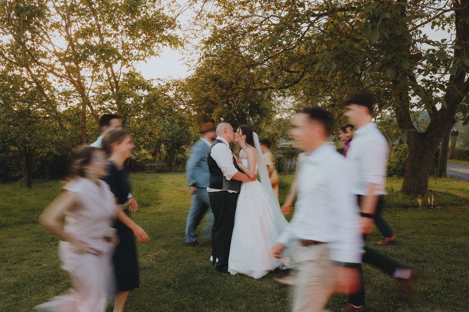 Bride and groom share a kiss surrounded by wedding guests on a lush green lawn during golden hour.