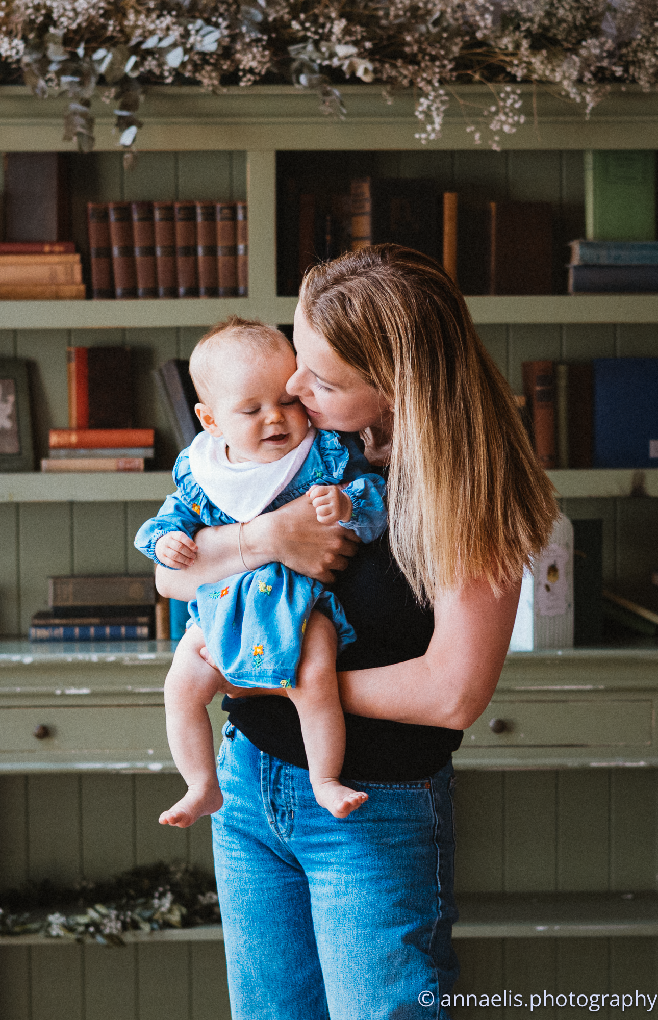 Parent and baby share sweet moment in front of bookshelf with spring blossoms.