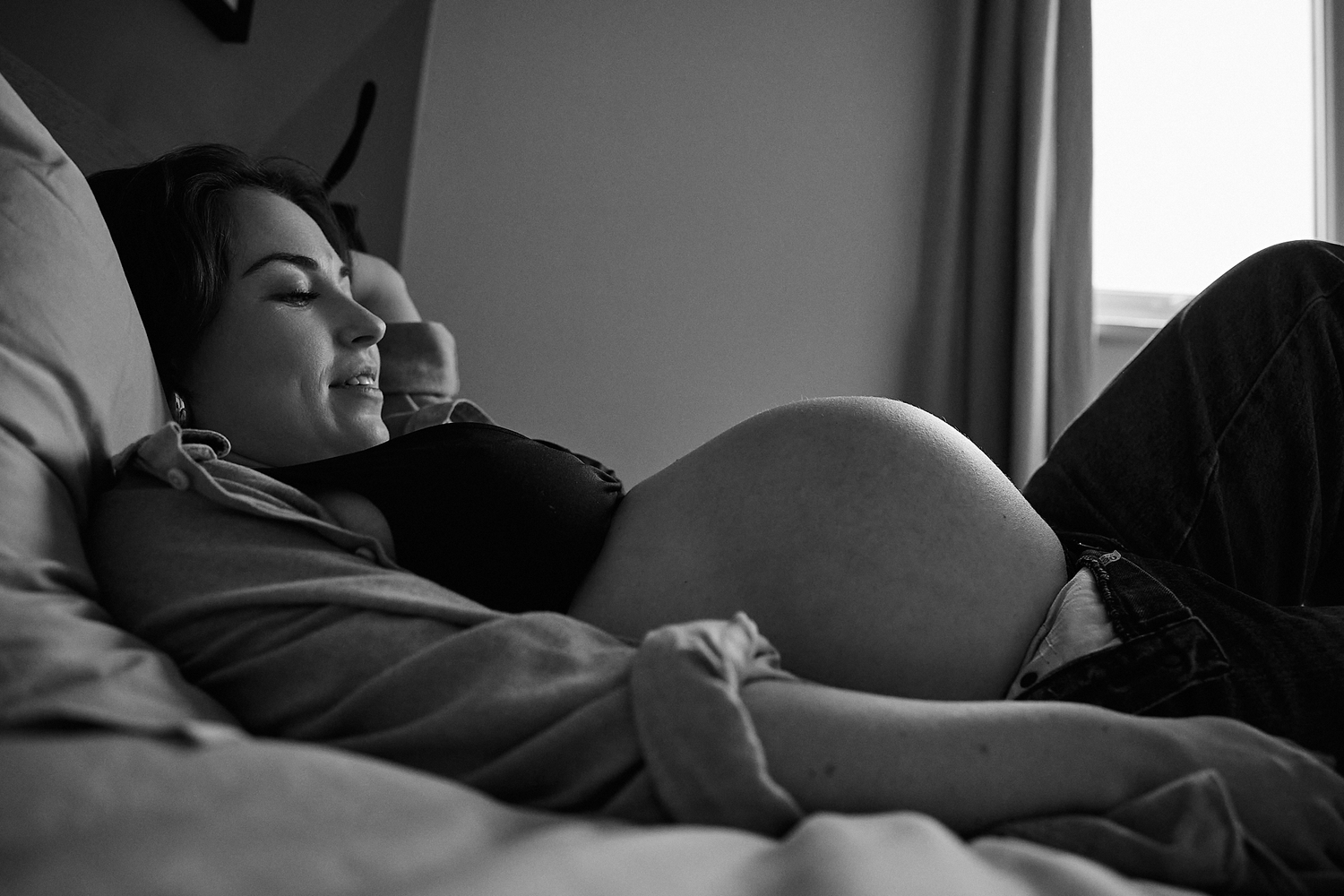 Black and white photo of a pregnant woman resting on a bed, cradling her baby bump with a serene smile.