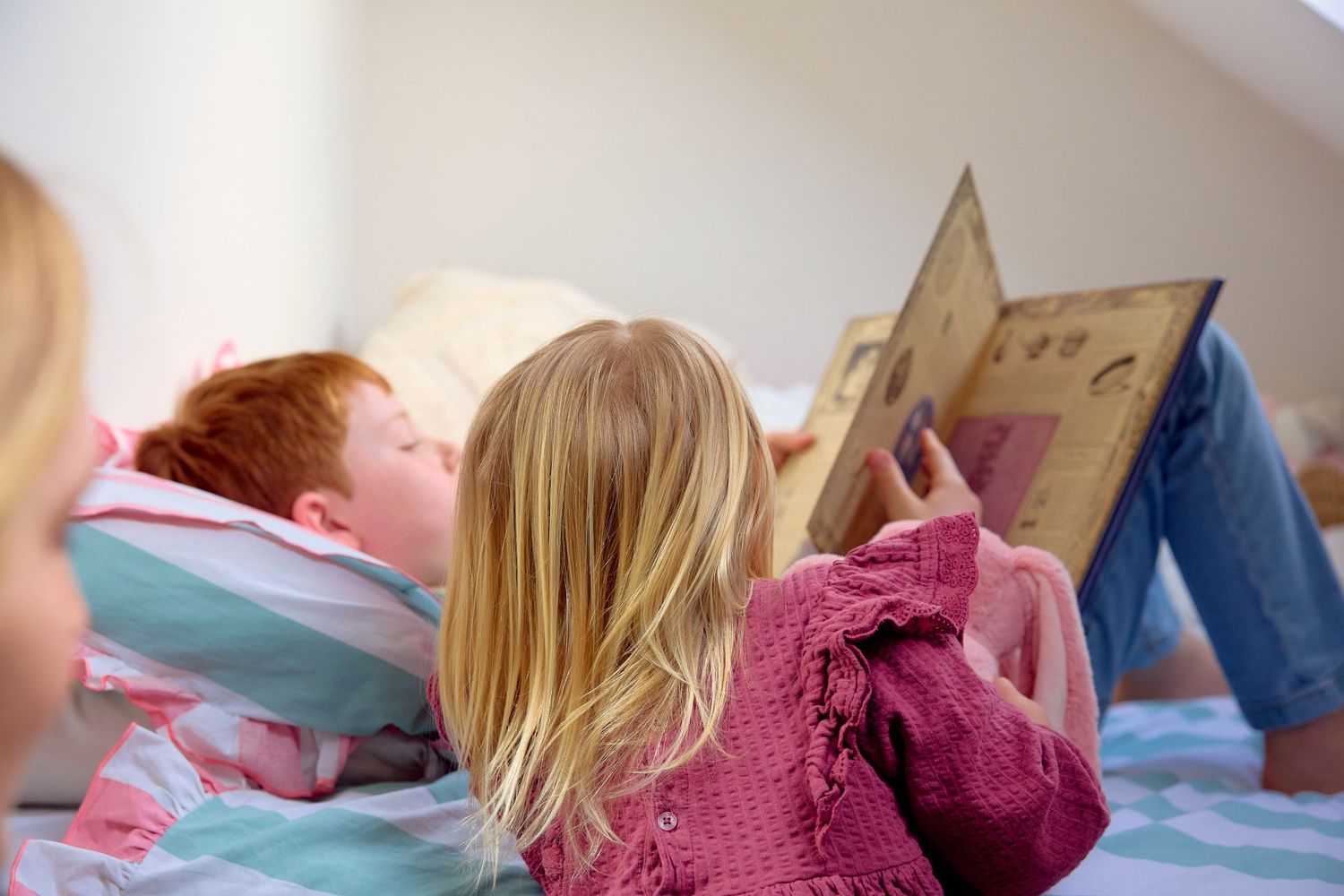 Two children lying on a bed reading a large book together, with pink pillows and a light blue bedspread.