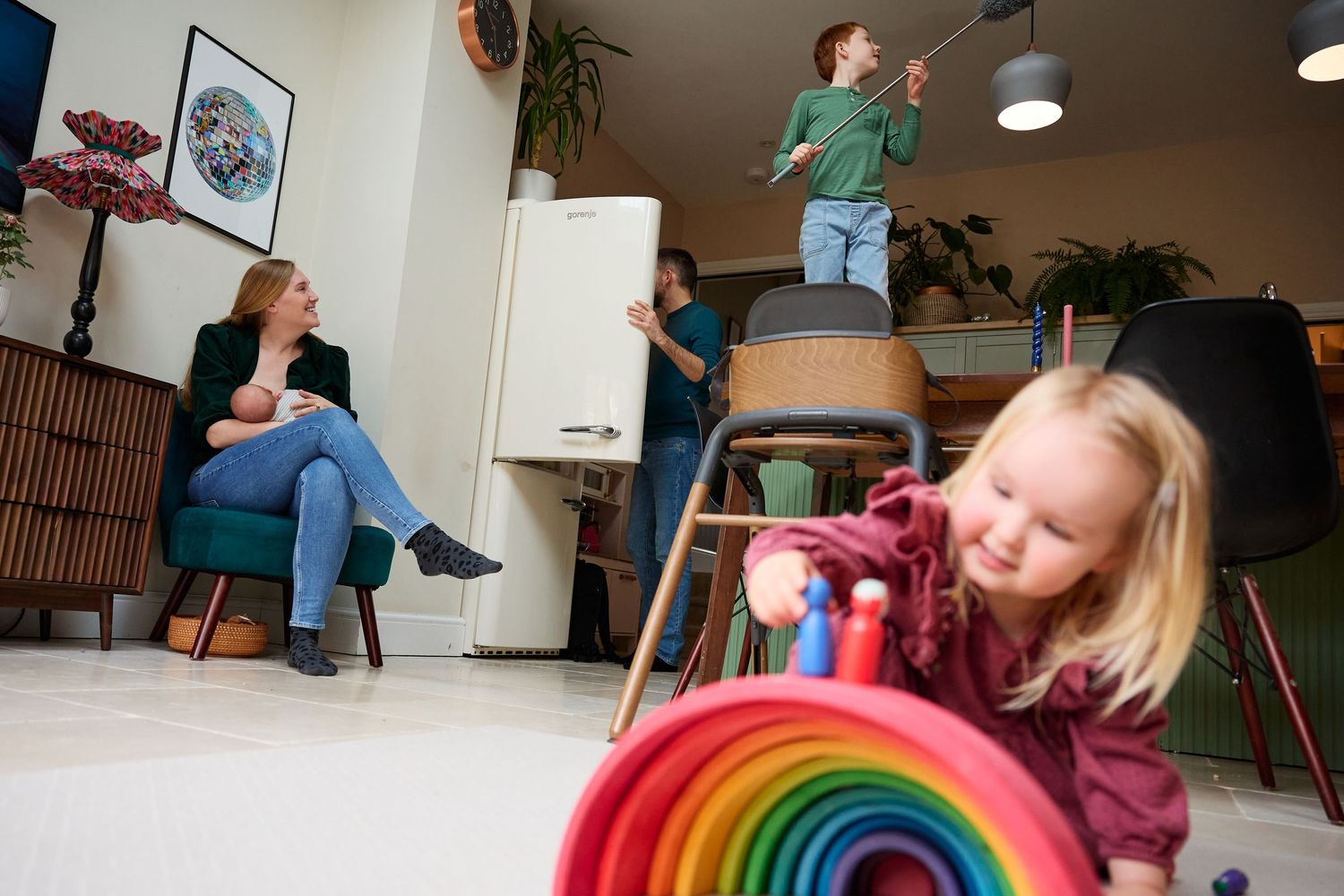 Young girl plays with colorful rainbow toy while mother relaxes and children play in bright modern living room.