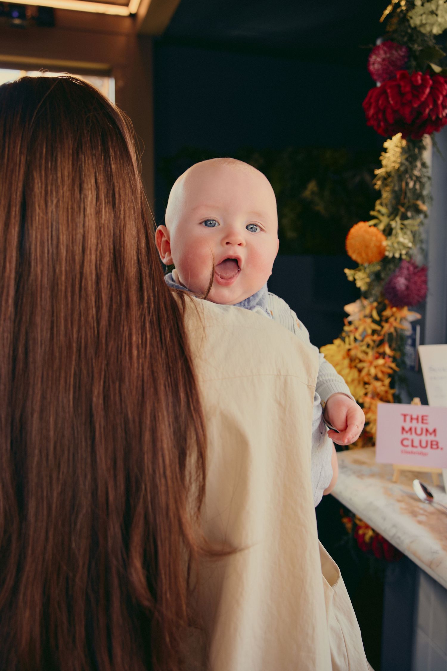 A mother holds her happy baby at The Mum Club event, surrounded by colorful flowers.