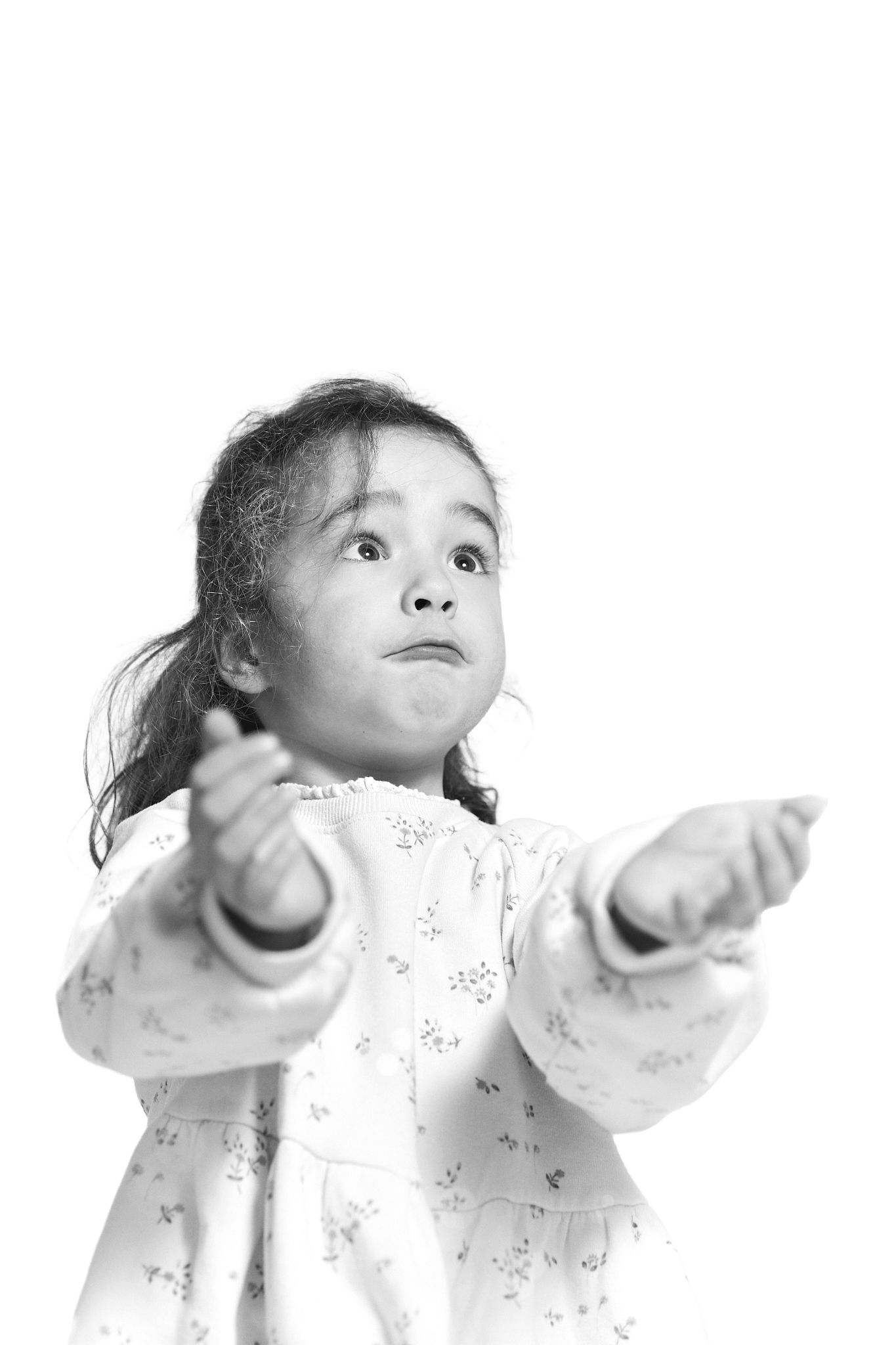 Black and white photo of a young girl in a floral dress reaching her arms out and looking up with curiosity.
