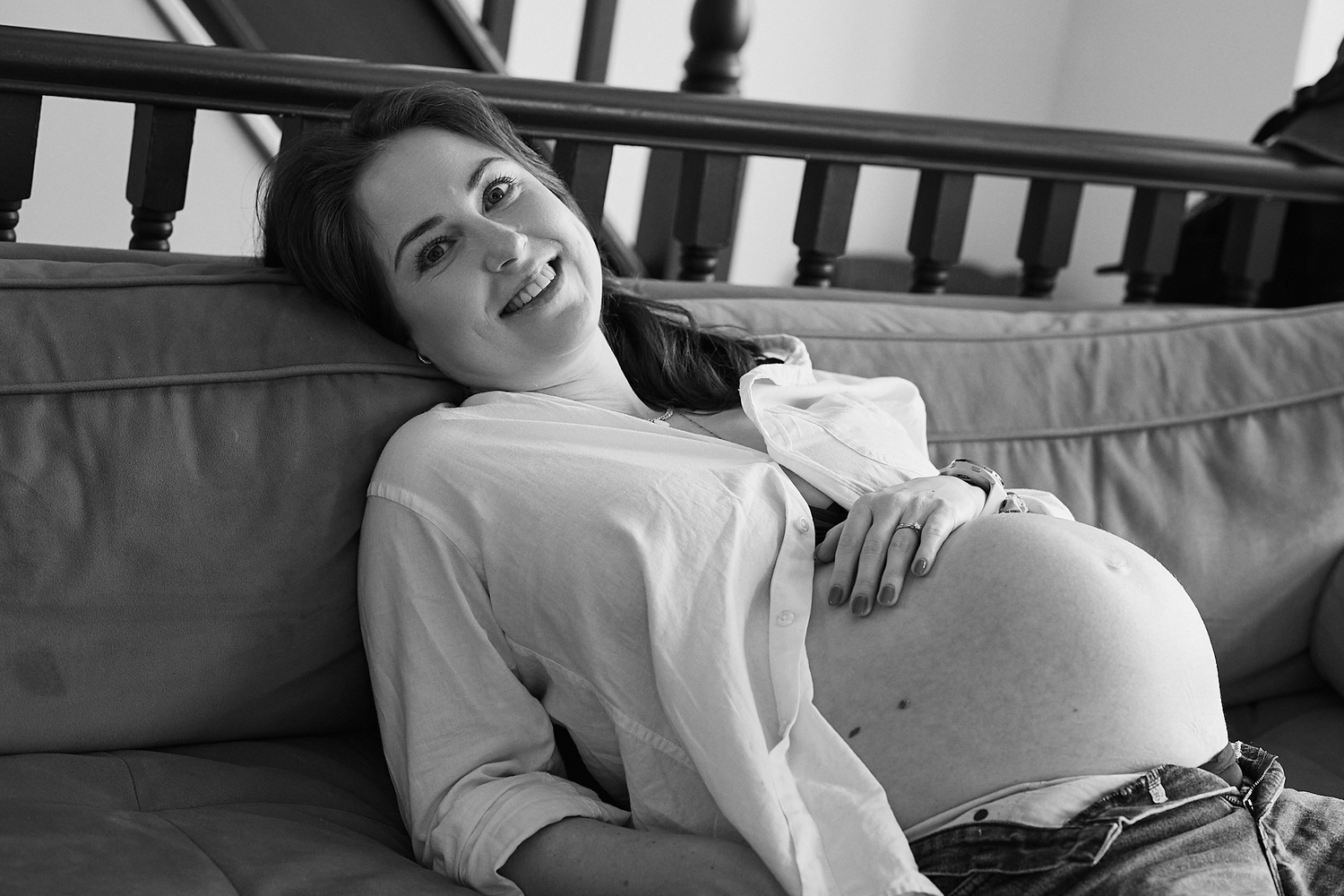 Smiling pregnant woman relaxing on a couch, cradling her baby bump, in a black and white maternity photo.