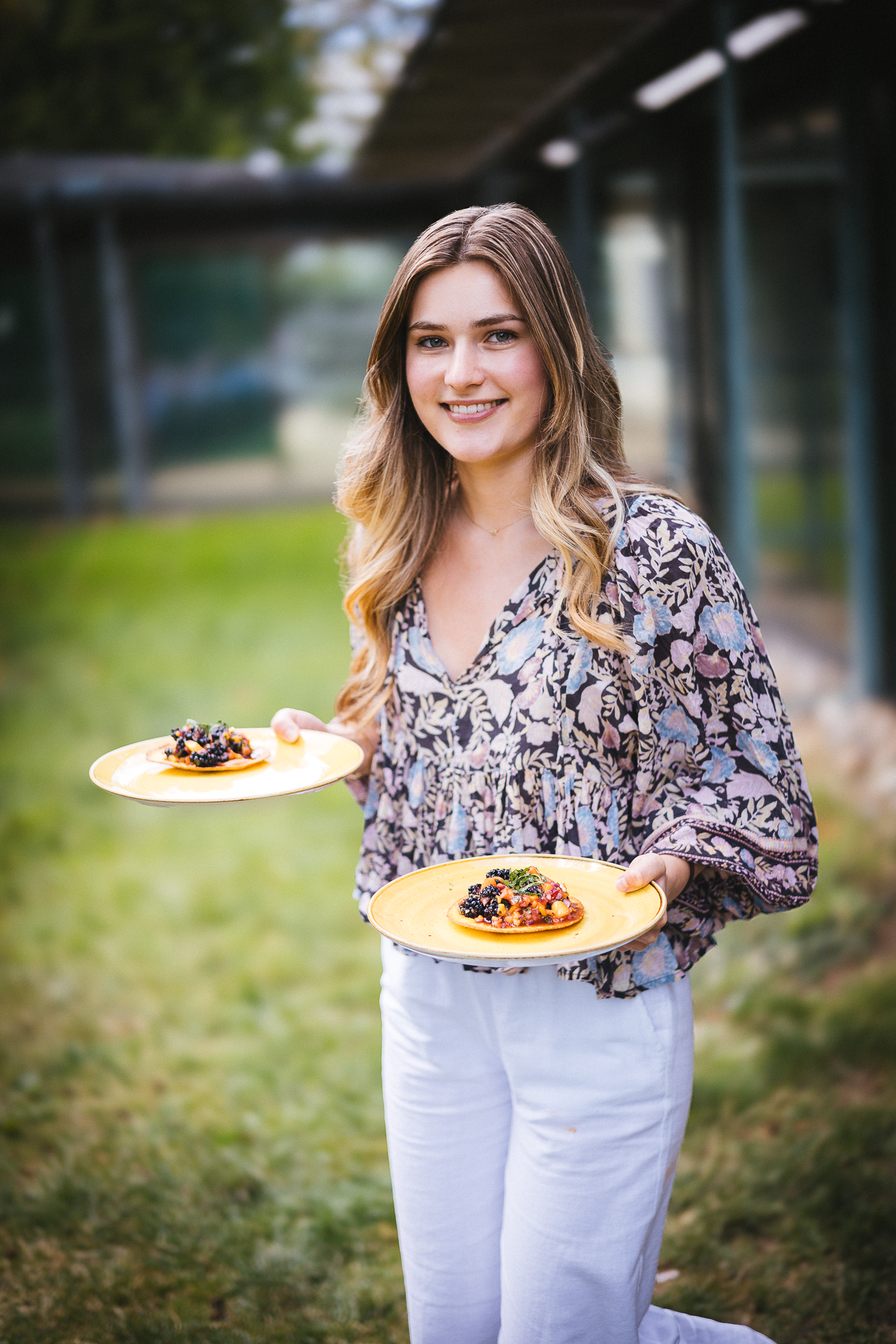 Person in floral print top and white pants holding yellow plates of colorful food outdoors.