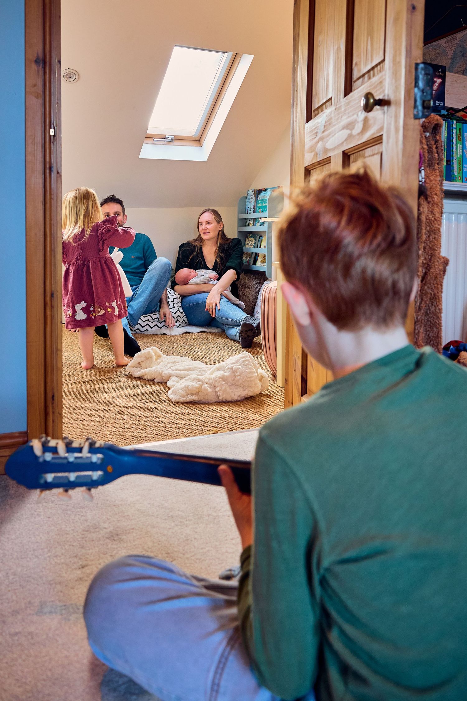 A boy plays guitar while his family relaxes in a cozy attic room with a newborn baby and young girl.