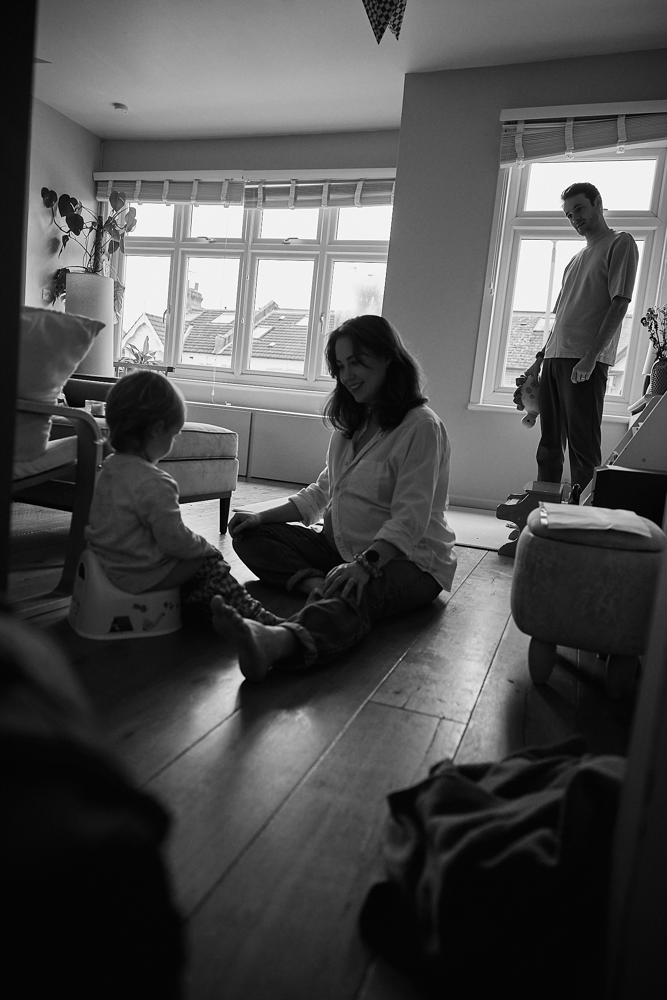 Black and white photo of a woman sitting on hardwood floor with toddler while man stands near bright windows.