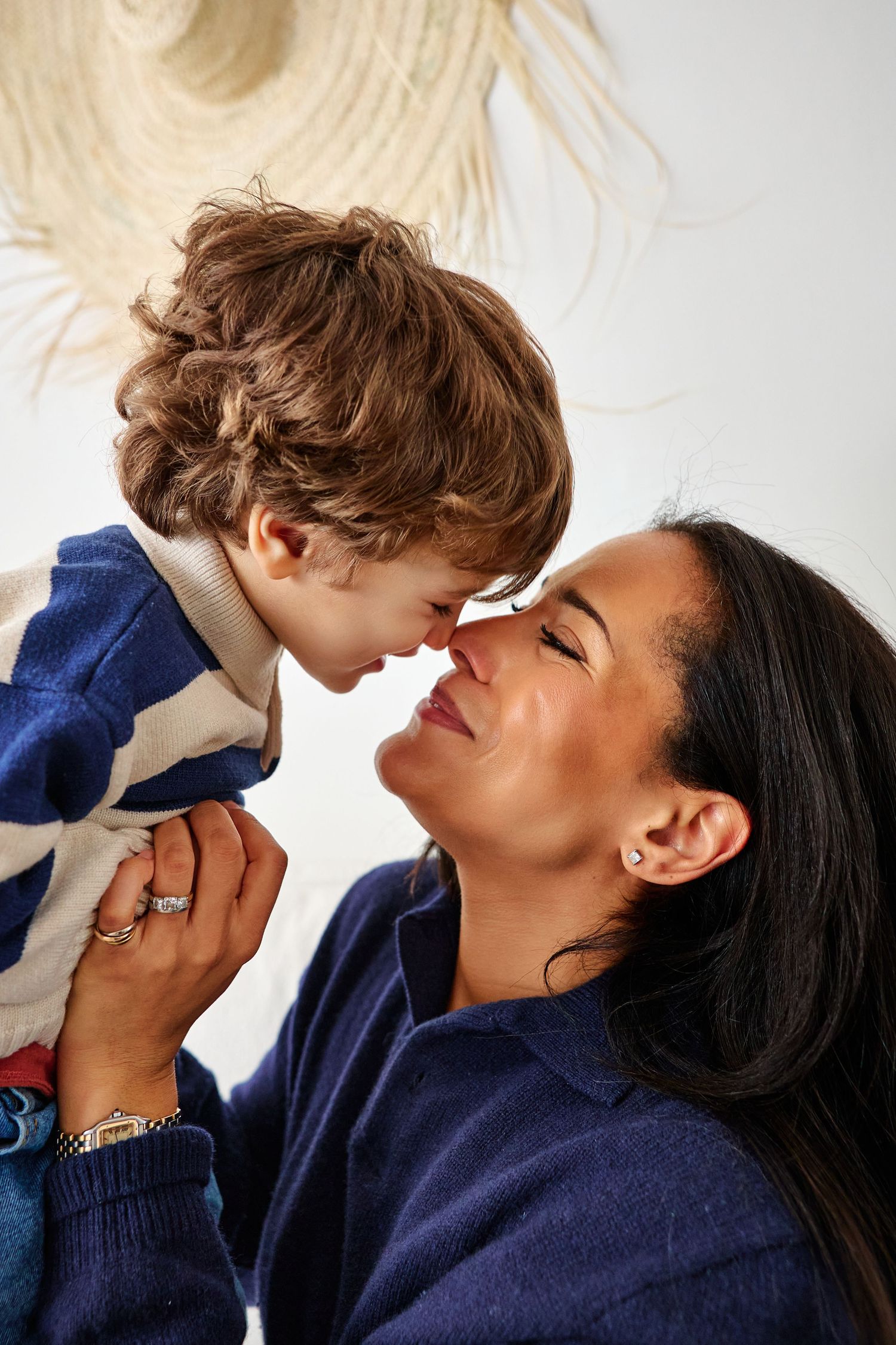 A mother and young boy share an Eskimo kiss, both smiling, dressed in blue sweaters against a white wall.