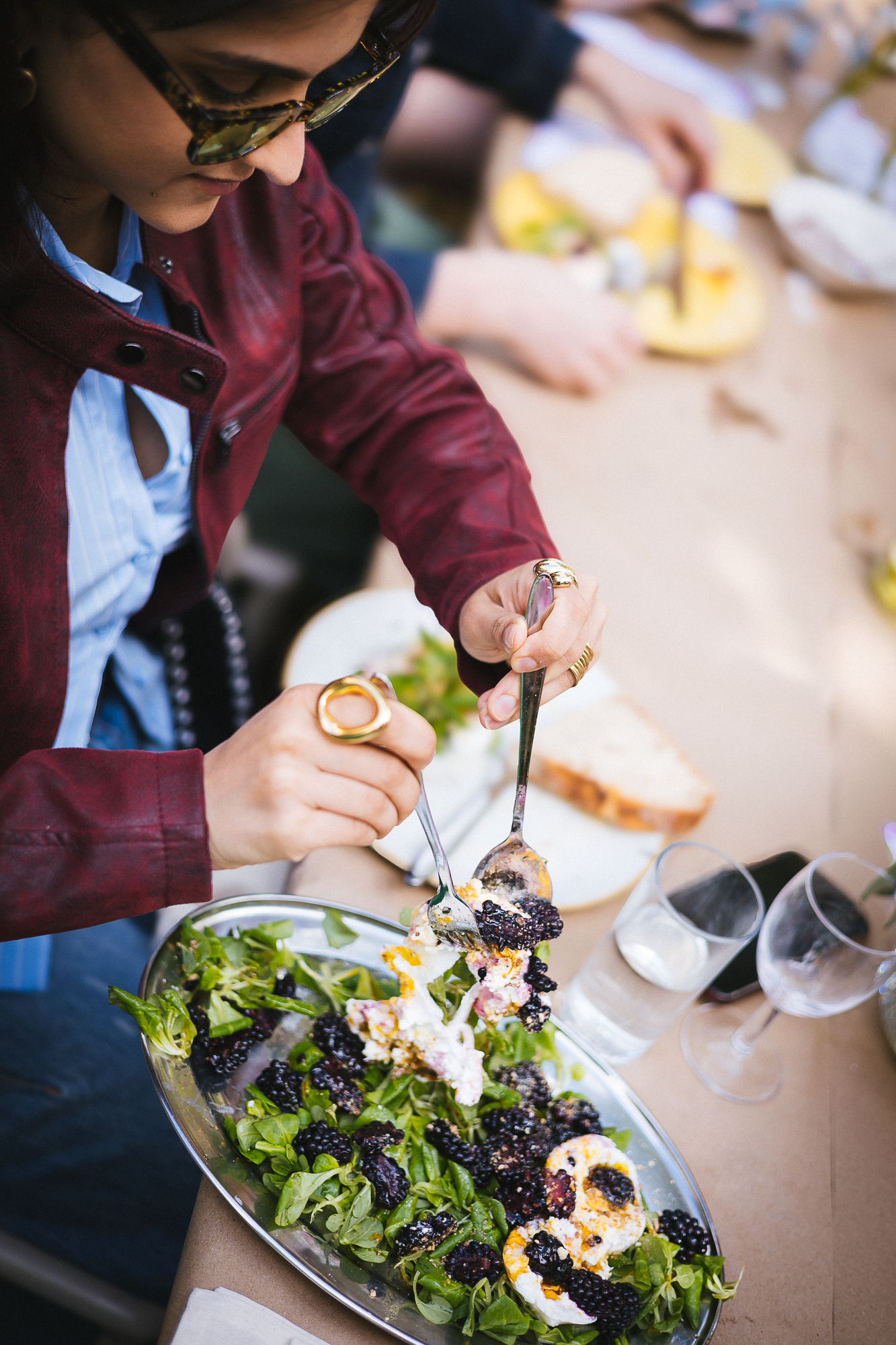 Close-up of hands serving fresh salad with mixed greens and toppings at an outdoor dining table.