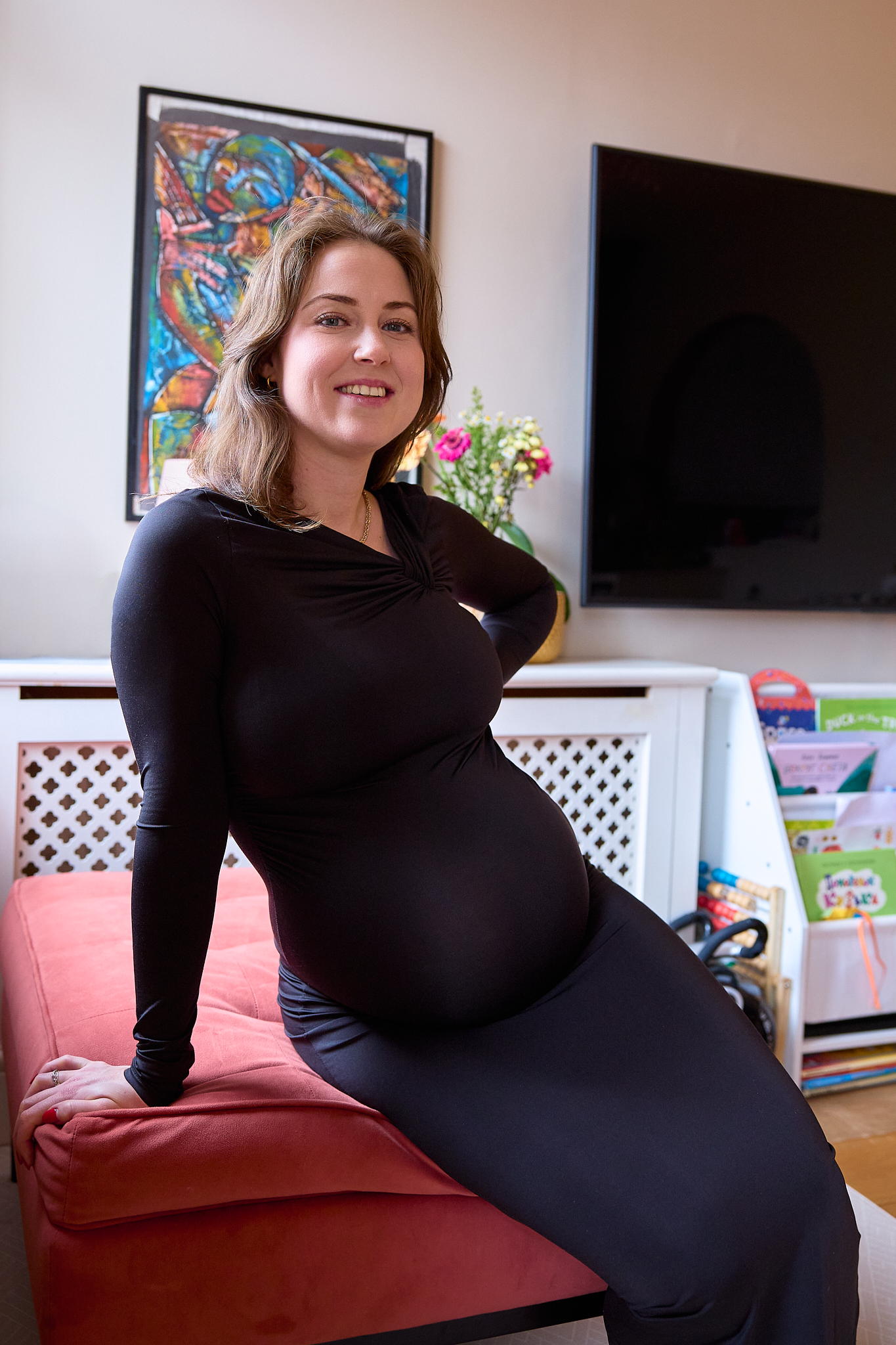 A person wearing a black long-sleeved top poses on a red couch in a living room with artwork on the walls.