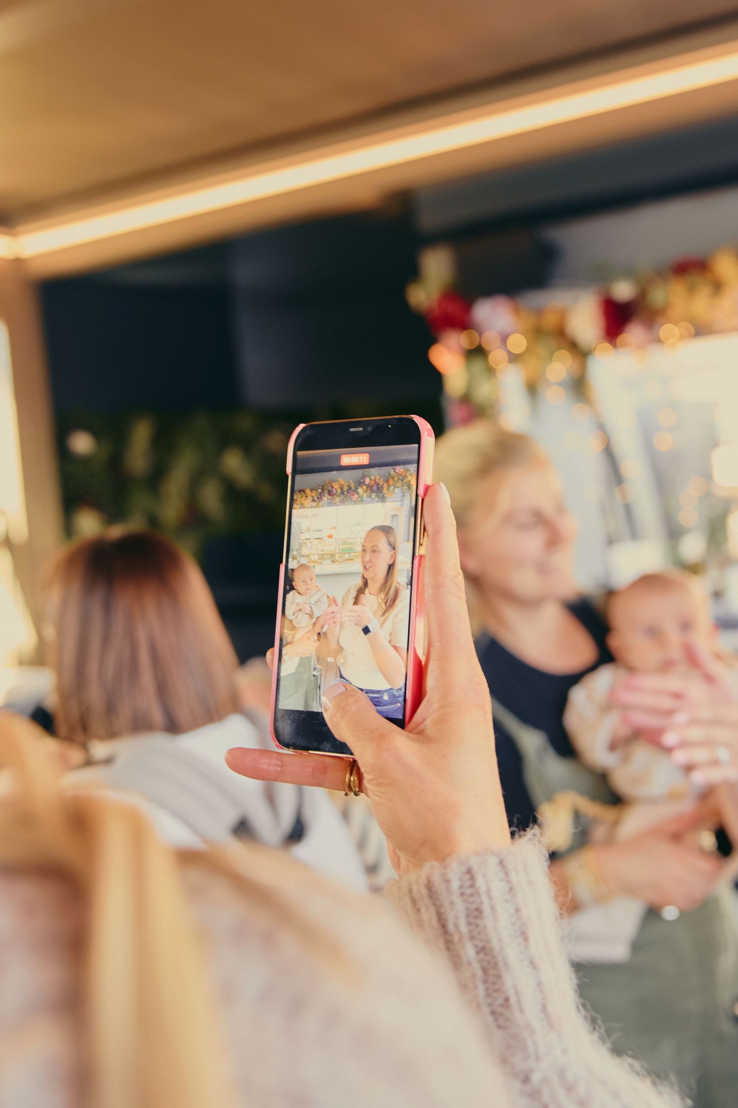 Woman photographs two mothers and a baby at a festive holiday market stall.