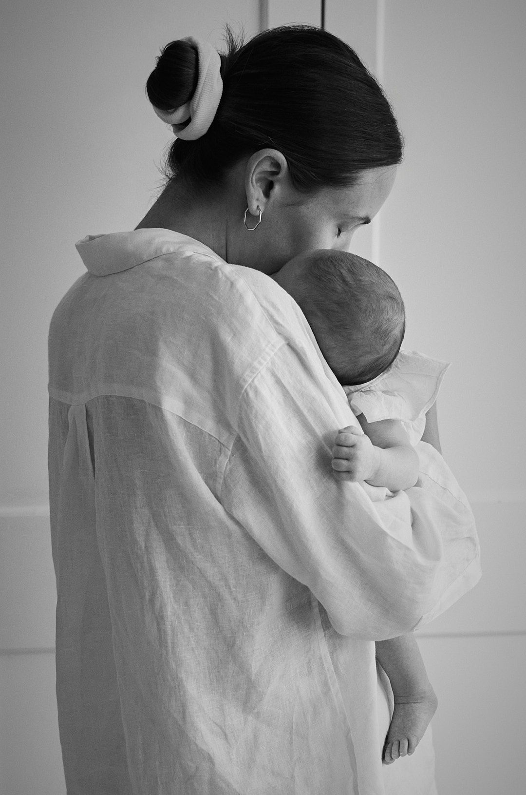Black and white photo of a mother in a white linen shirt tenderly holding and kissing her newborn baby.