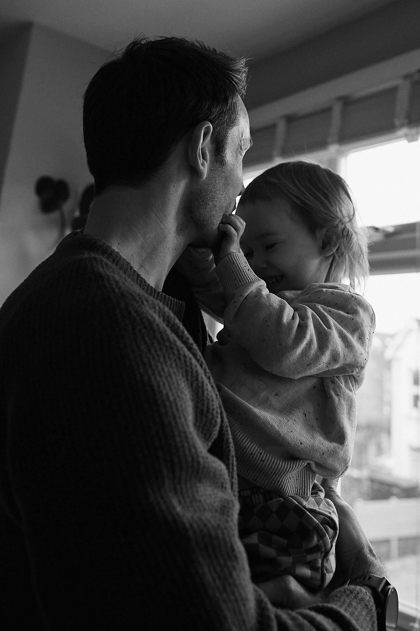 Black and white photo of a father holding his toddler child near a window in a tender moment.