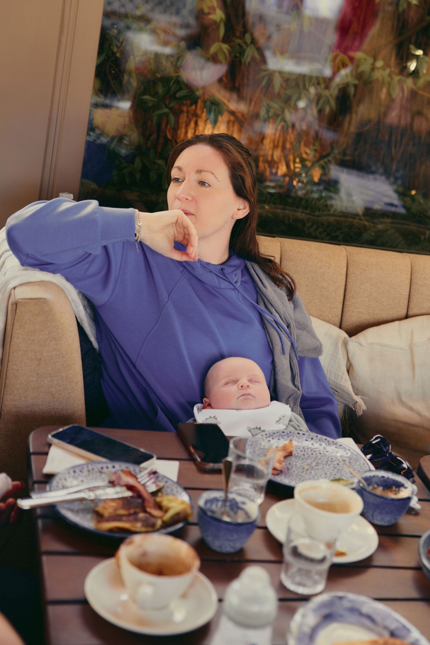 A tired mother in a blue sweater sits in a cafe with her newborn baby, enjoying coffee and food.