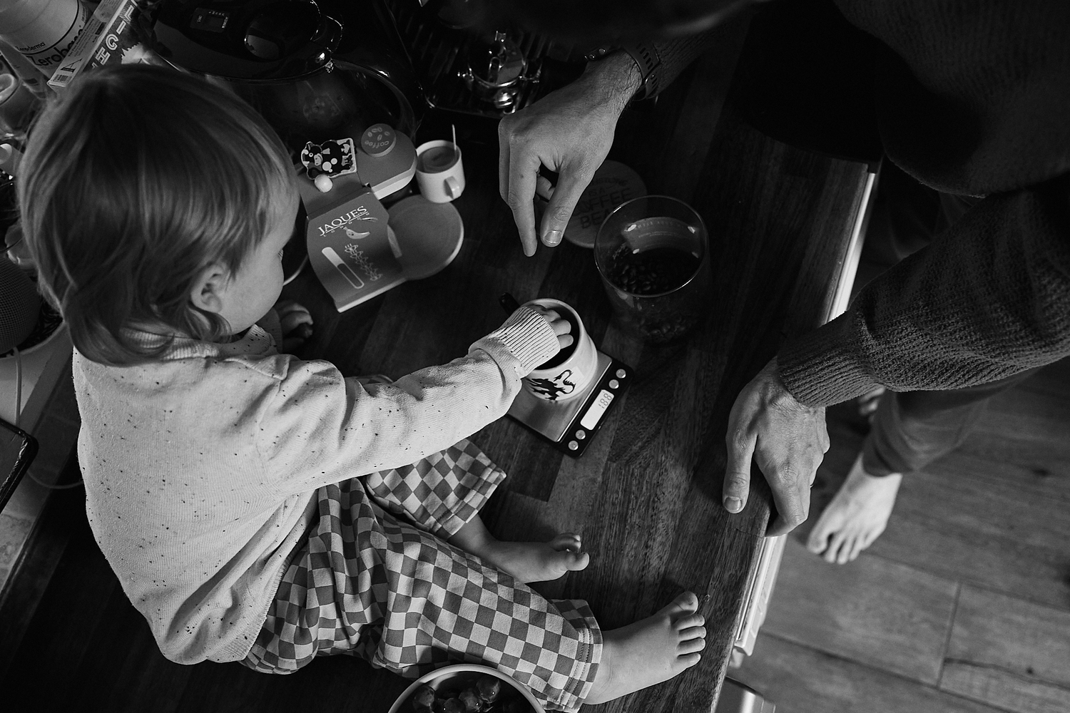 Black and white photo of a child and adult sitting at a table, sharing a moment over drinks and a small device.
