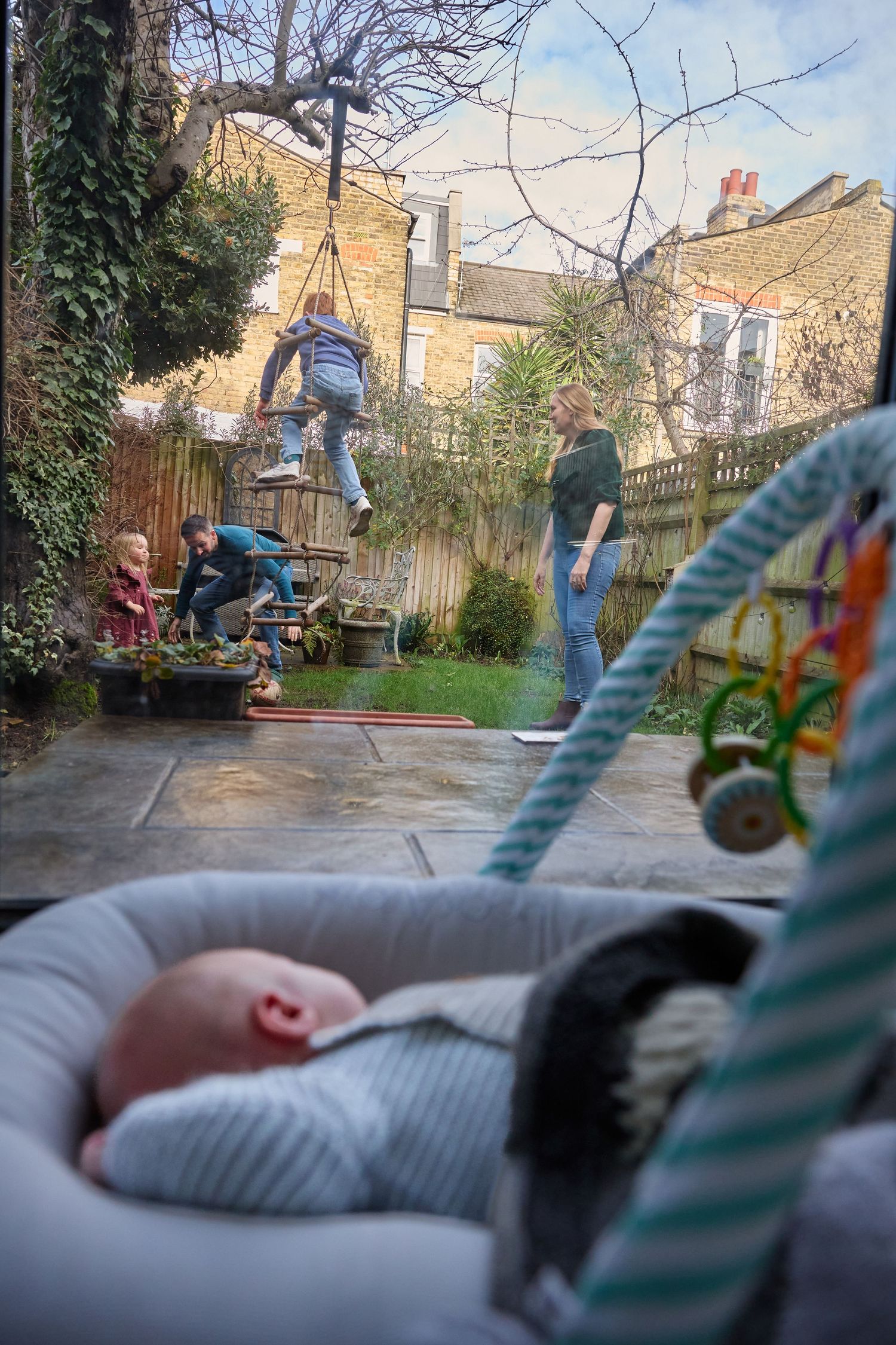 Baby sleeping in bassinet indoors while family plays in garden with child on rope swing.